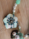 A close-up of a handmade beaded necklace or ornament featuring a large iridescent flower charm with blue rhinestone accents, white faux pearls, and light green beads, laid on a light wood surface.