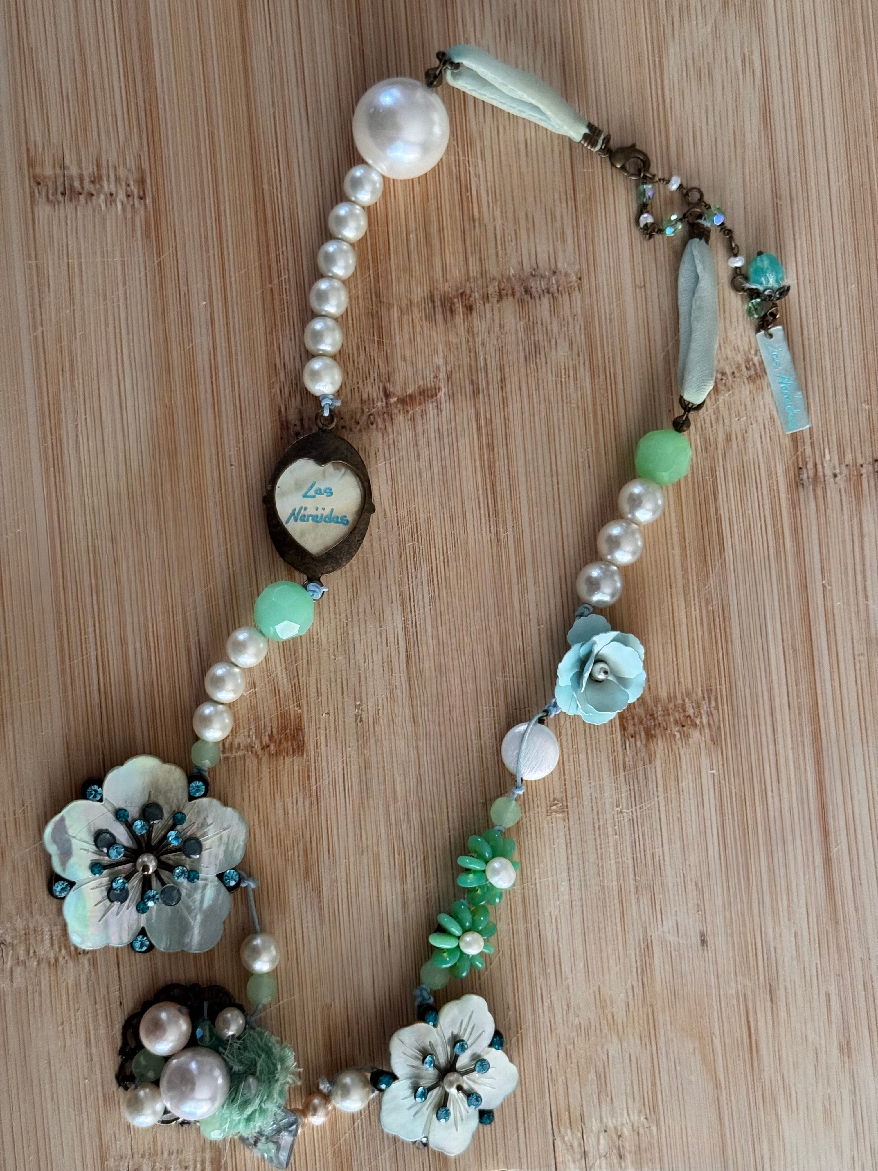 A handmade beaded necklace with faux pearls, light blue and mint green beads, flower charms, and a central heart-shaped pendant, displayed on a light wood background.