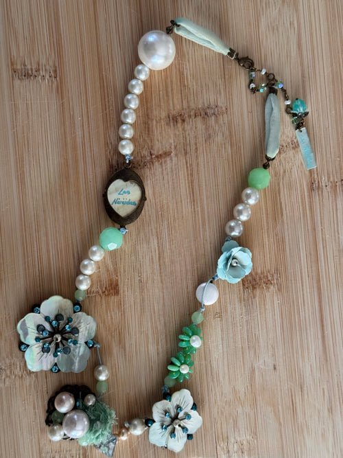 A handmade beaded necklace with faux pearls, light blue and mint green beads, flower charms, and a central heart-shaped pendant, displayed on a light wood background.