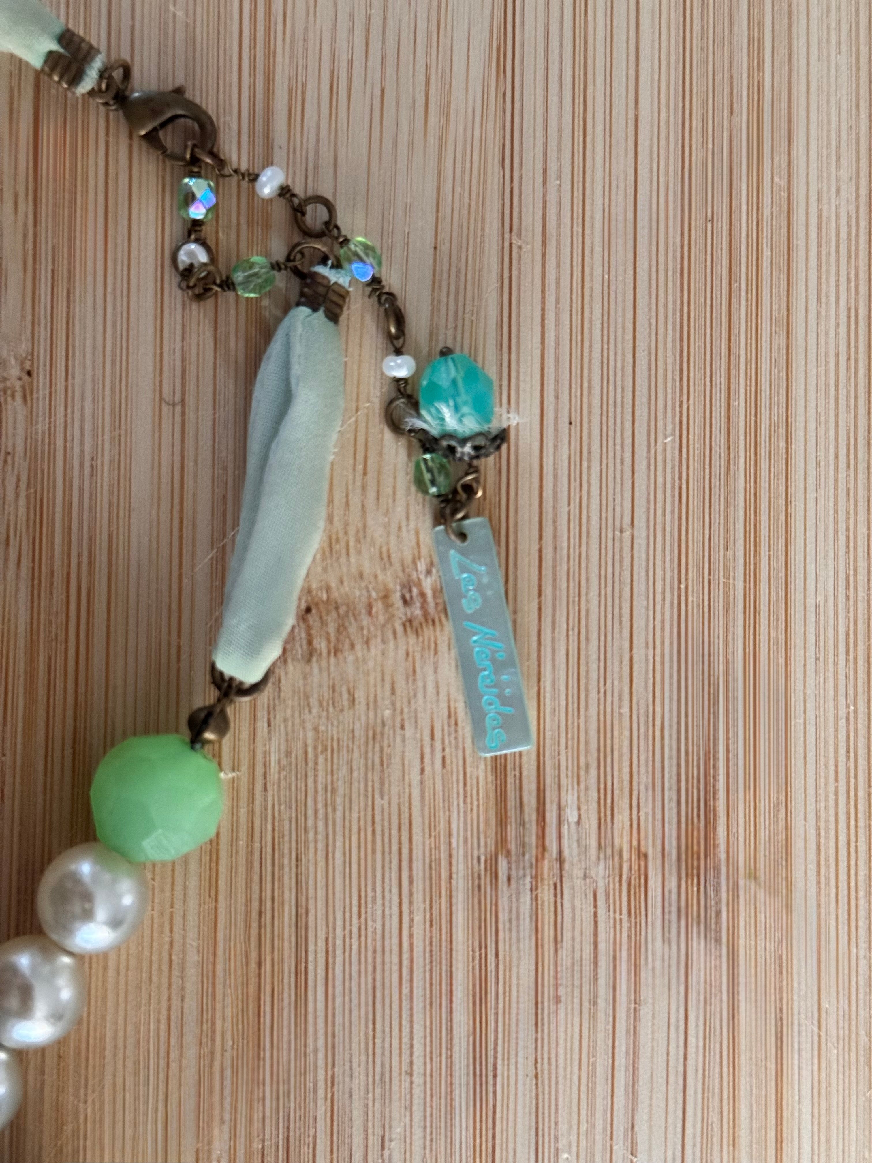 A close-up of a handmade beaded necklace laid flat on a light wood grain surface. The piece features a light blue-green glass fragment, a round light green bead, white faux pearls, small iridescent beads, and a small engraved metal tag of the designer, This image shows the clasp of the necklace.