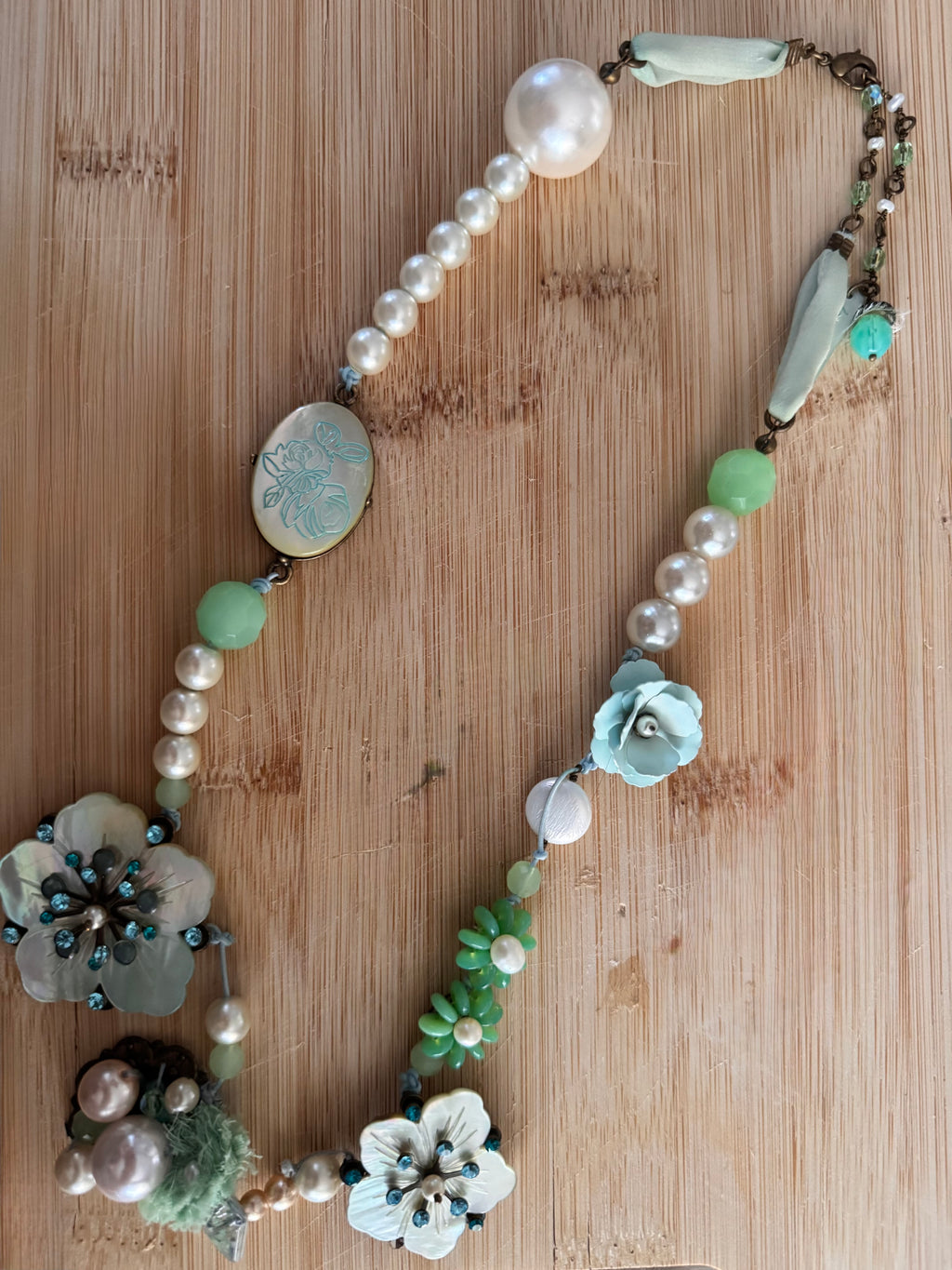 A handmade beaded necklace with faux pearls, mint green beads, light blue flower charms, and a carved oval pendant, displayed on a light wood background.