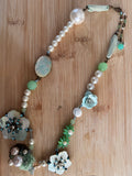 A handmade beaded necklace with faux pearls, mint green beads, light blue flower charms, and a carved oval pendant, displayed on a light wood background.