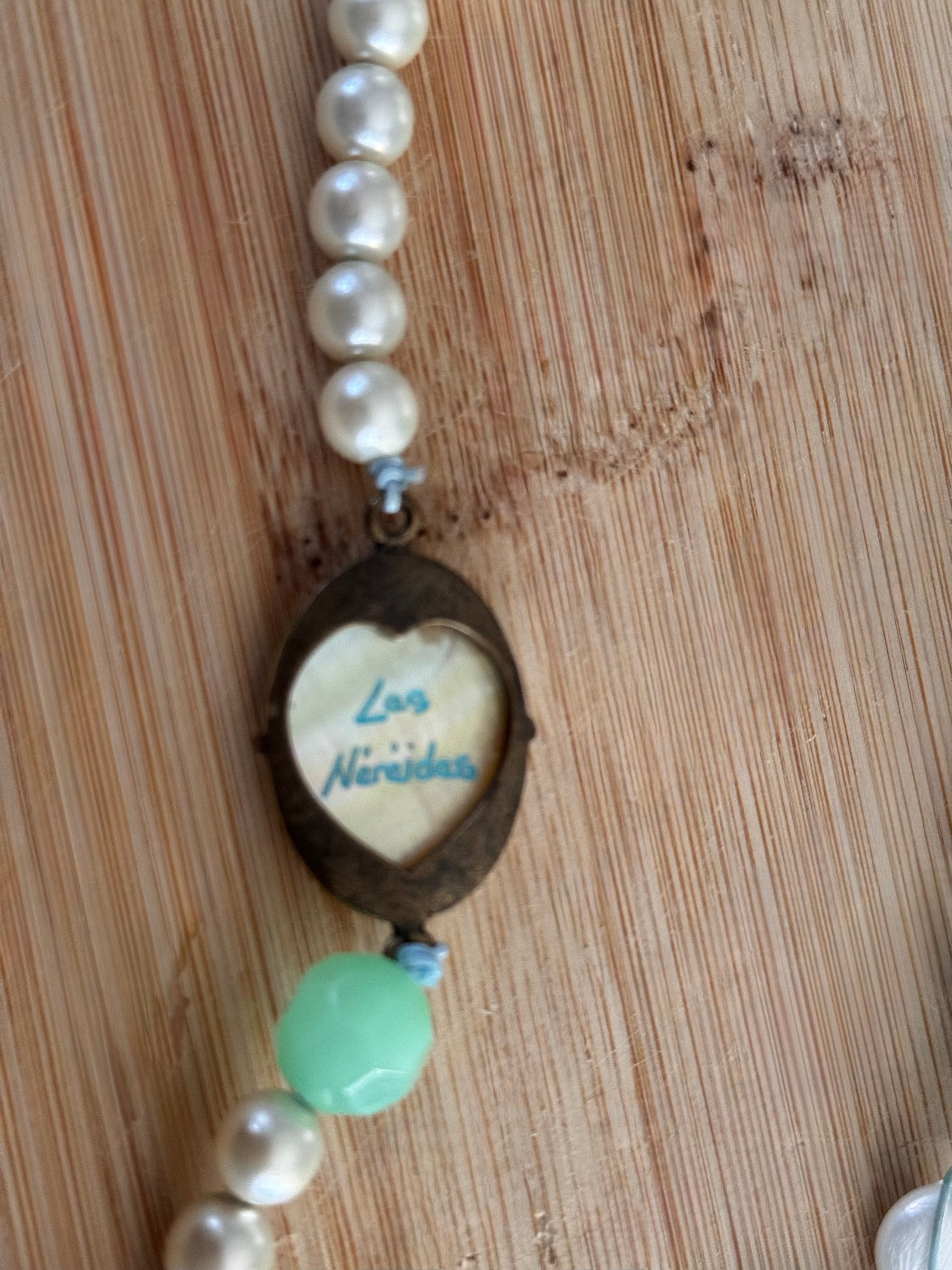 A close-up of a handmade beaded necklace featuring white faux pearls, a dark metal heart-shaped frame with a small white insert reading 'Les Nereides' in blue script, a light green bead, and a light blue accent bead, laid flat on a light wood grain surface.