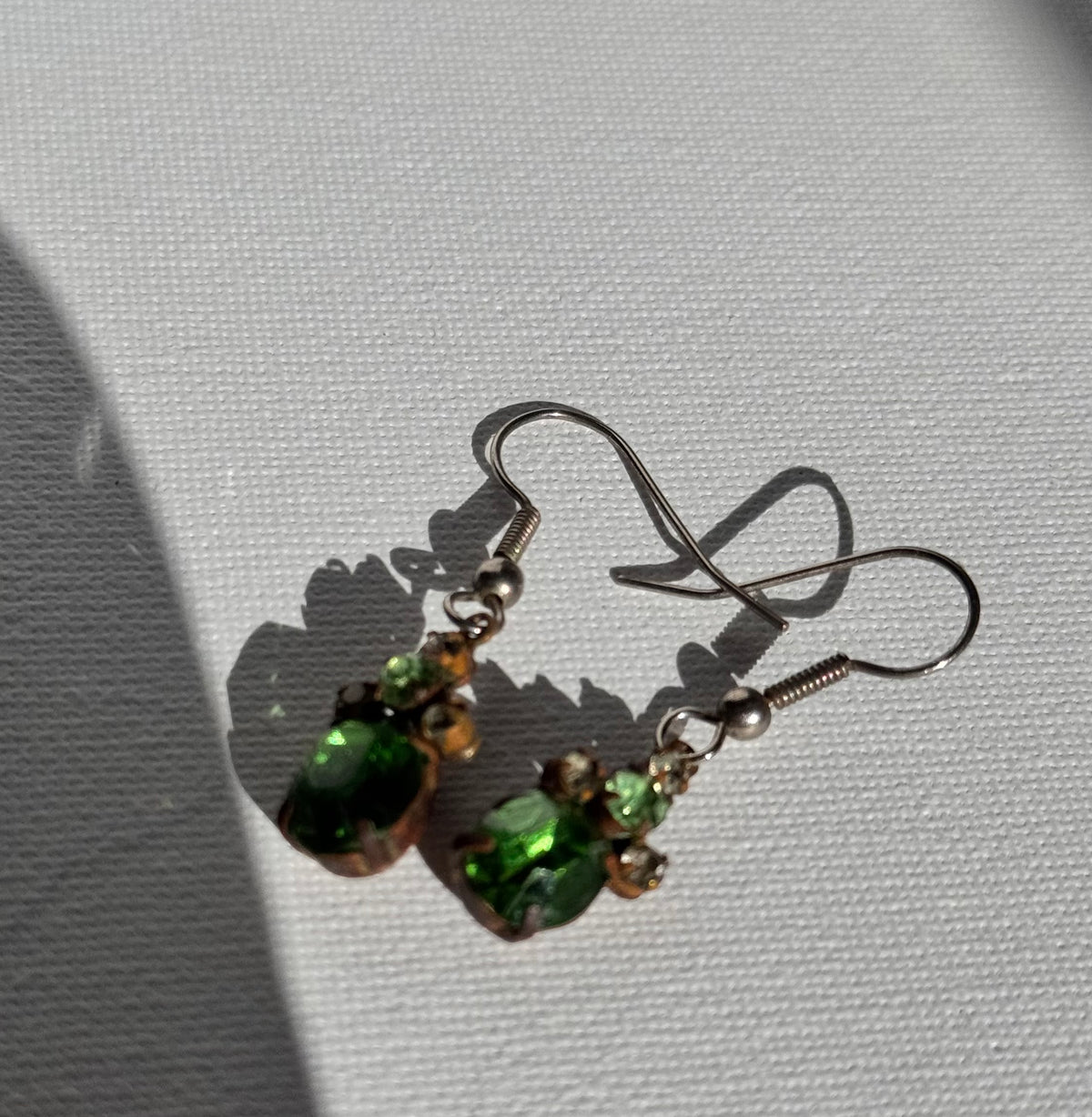 A pair of handmade dangle earrings with green faceted beads, small metallic beads, and silver fishhook wires, displayed on a textured white surface with a soft shadow.
