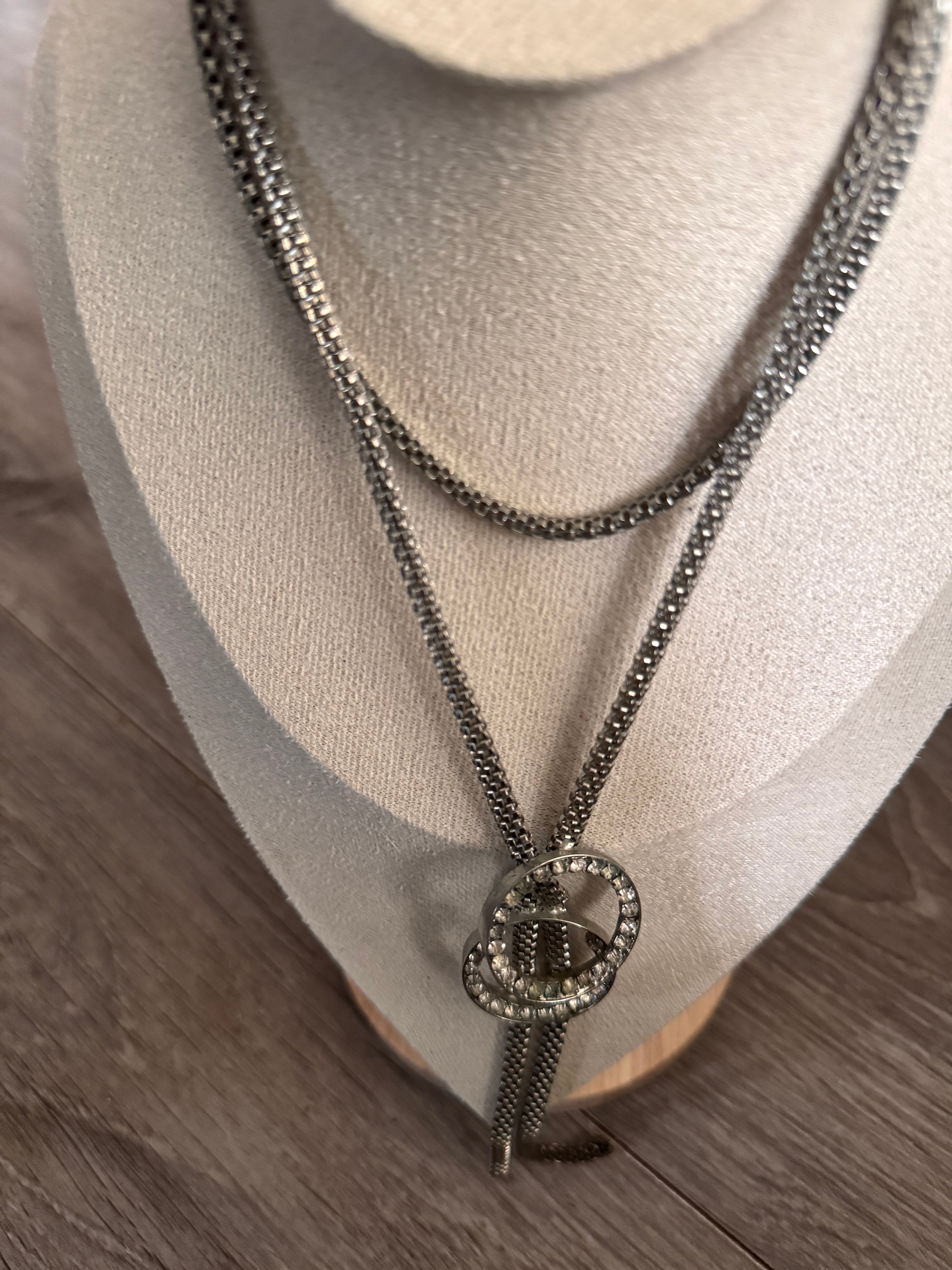 A silver-toned double-strand chain necklace with a circular crystal-encrusted pendant, displayed on a beige jewelry bust over a wood-grain surface.