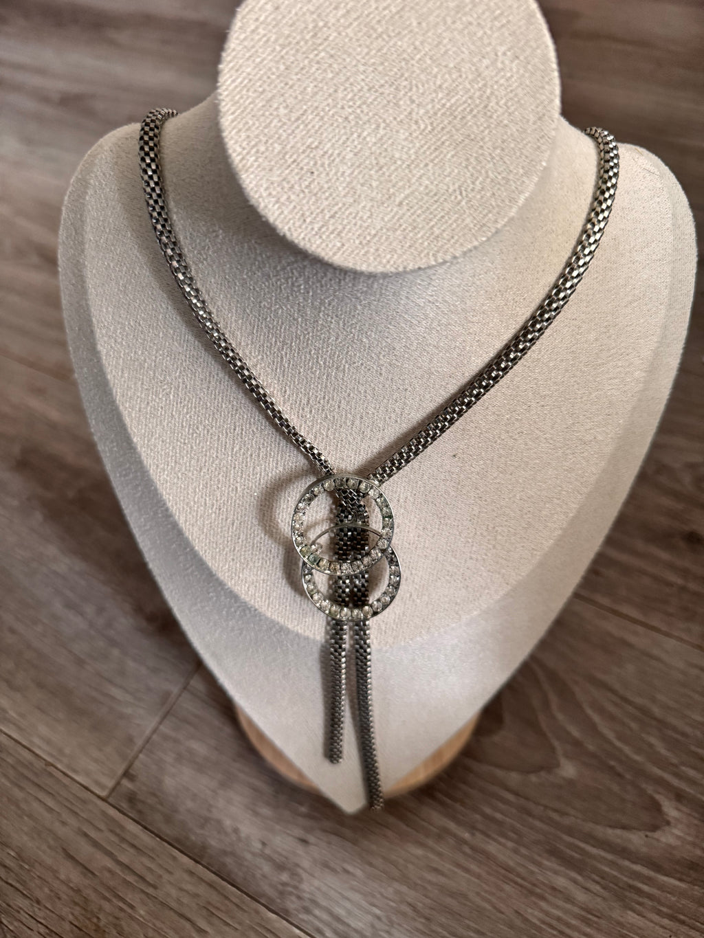 A silver-toned chain necklace with a decorative interlocking double-ring centerpiece, displayed on a cream-colored jewelry bust over a wood floor background.