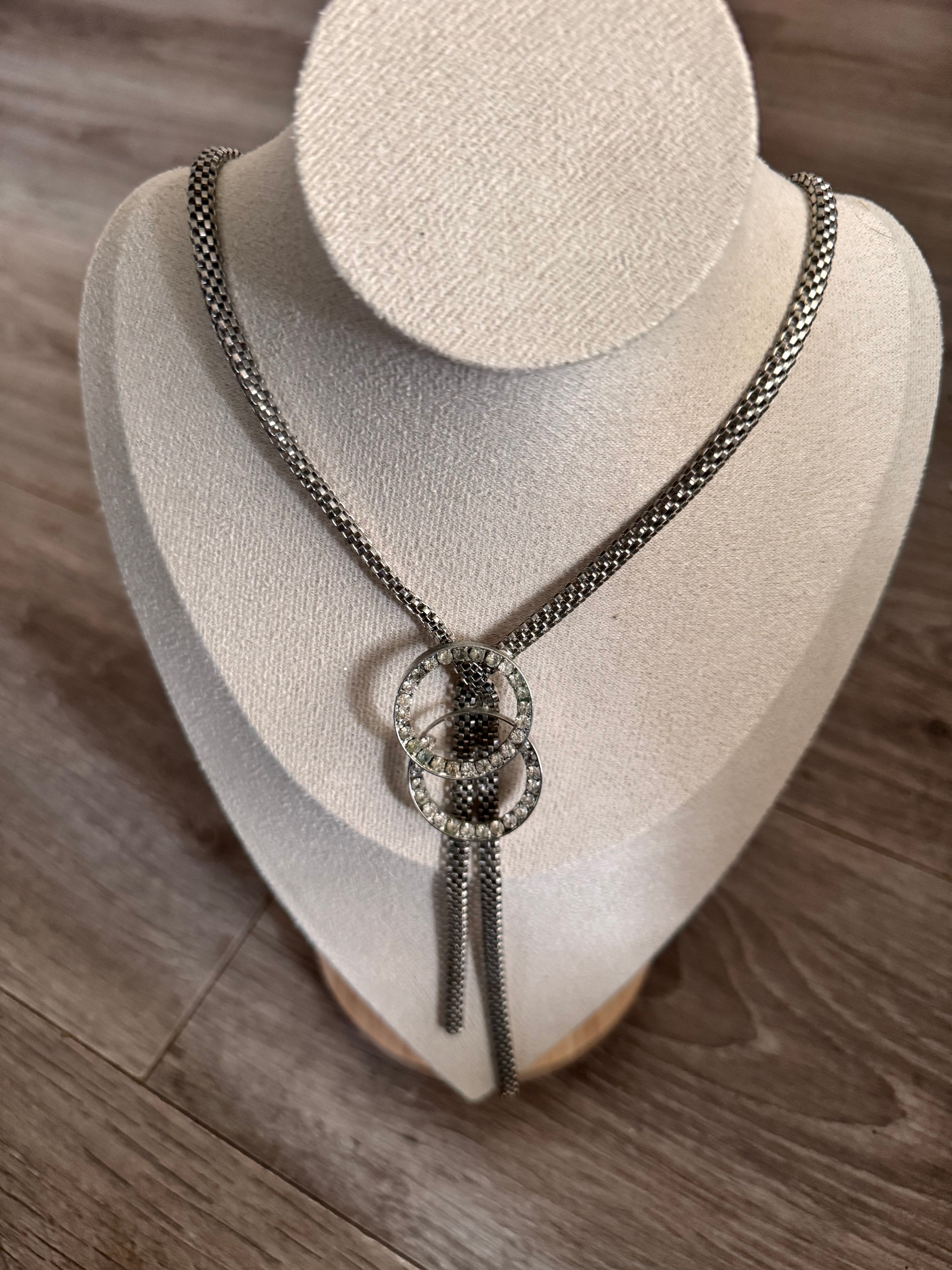 A silver-toned chain necklace with a decorative interlocking double-ring centerpiece, displayed on a cream-colored jewelry bust over a wood floor background.