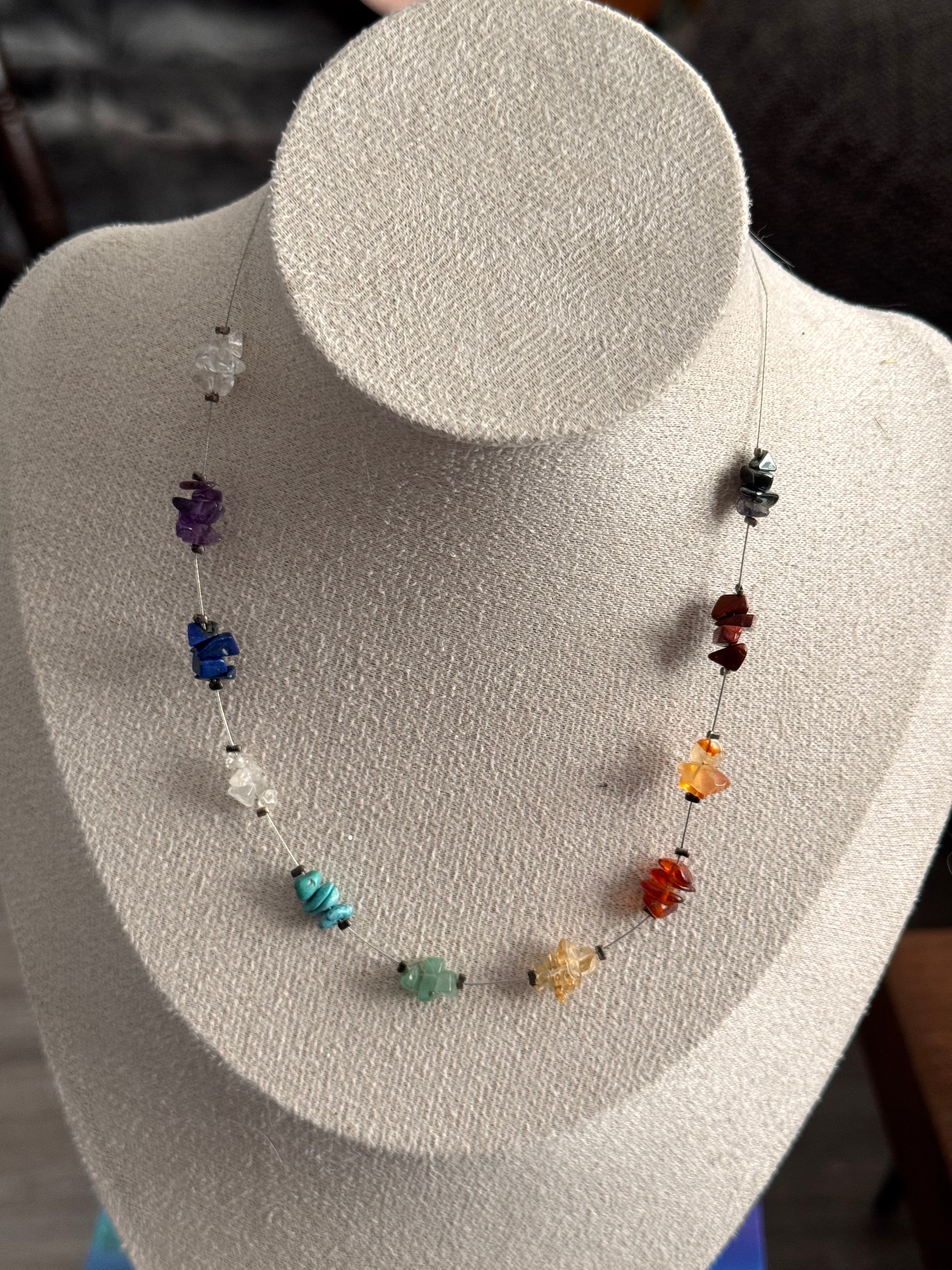 A beaded crystal necklace displayed on a white textured jewelry bust. The necklace features multiple small, polished gemstone beads in various colours including clear crystal, purple amethyst, red & blue agate, green possibly jade, yellow sunburst quartz, orange quartz, strawberry or melon quartz and brown agate type, strung on a thin metal cord chain.