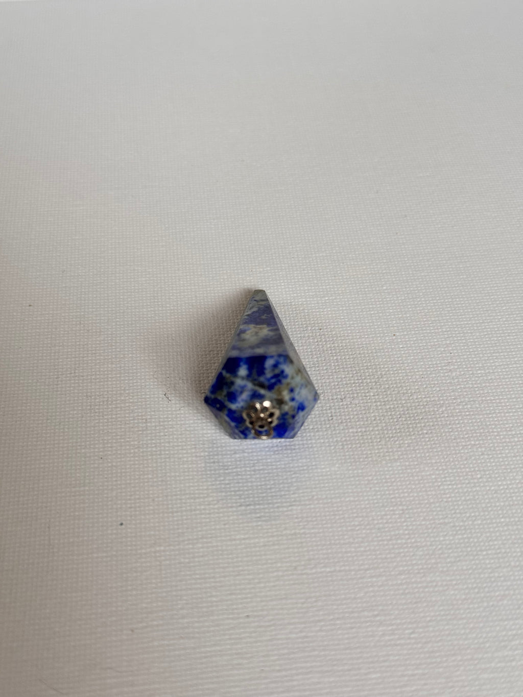 A small blue and white speckled pyramid-shaped crystal or gemstone with a silver metal loop at its center, placed on a white textured fabric background.