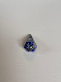 A small blue and white speckled pyramid-shaped crystal or gemstone with a silver metal loop at its center, placed on a white textured fabric background.