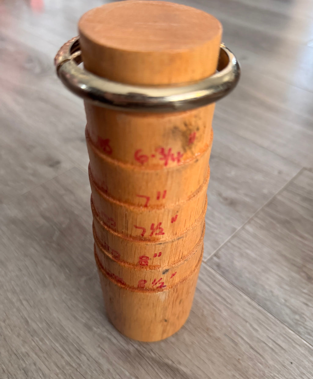 The reverse angle of the gold toned bracelet on a traditional wooden cylindrical mandrel. displays measurement markings the bracelet at the top, standing on a grey wood-look floor.