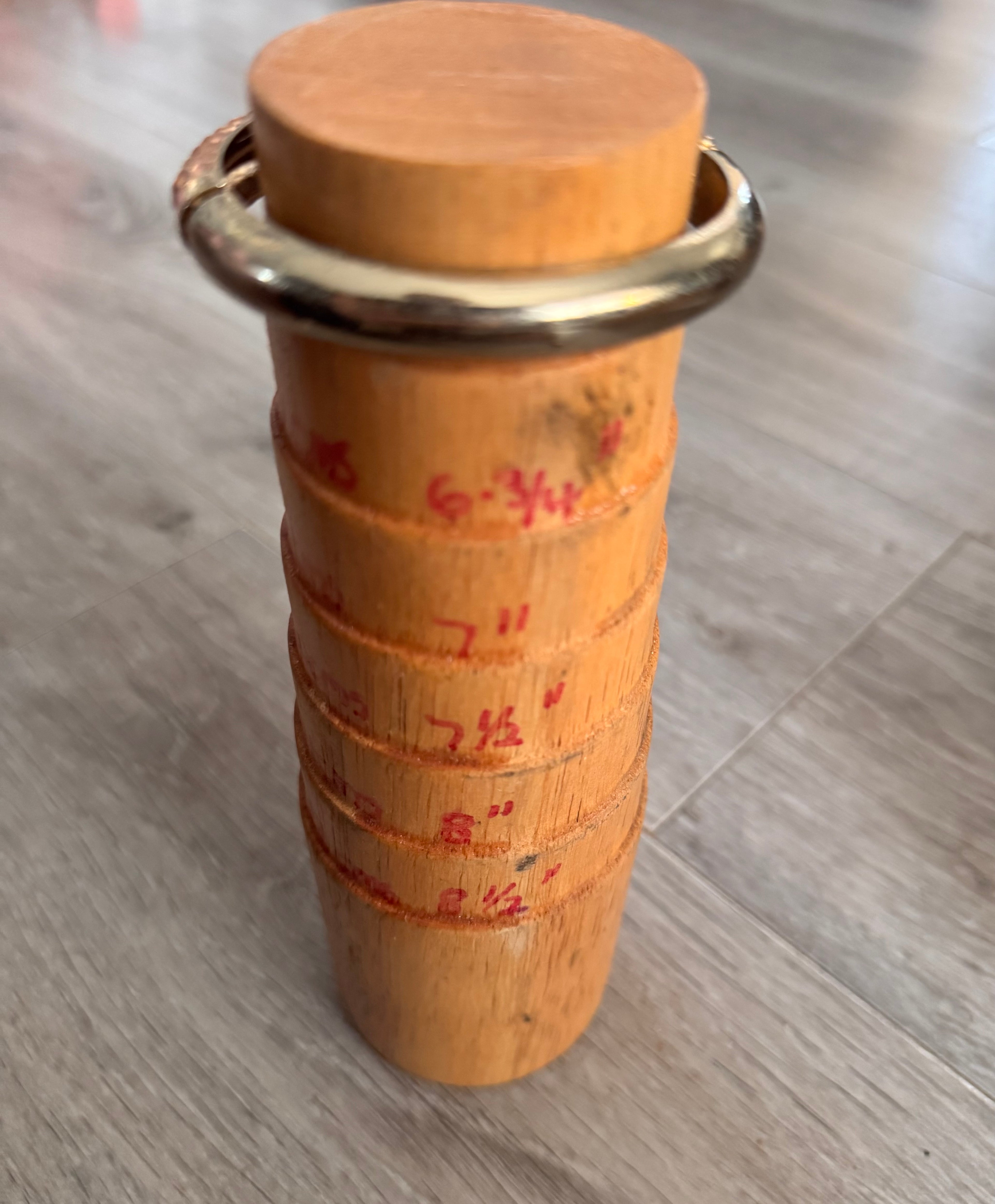 The reverse angle of the gold toned bracelet on a traditional wooden cylindrical mandrel. displays measurement markings the bracelet at the top, standing on a grey wood-look floor.