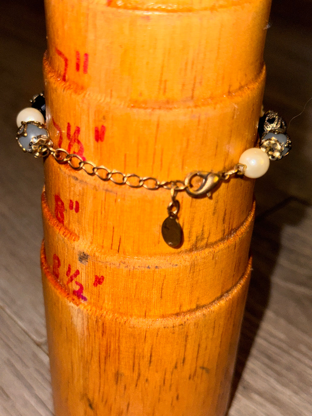 A gold charm bracelet by Accessorize with white pearl-like beads and dark metallic accents, displayed on a wooden cylindrical stand.