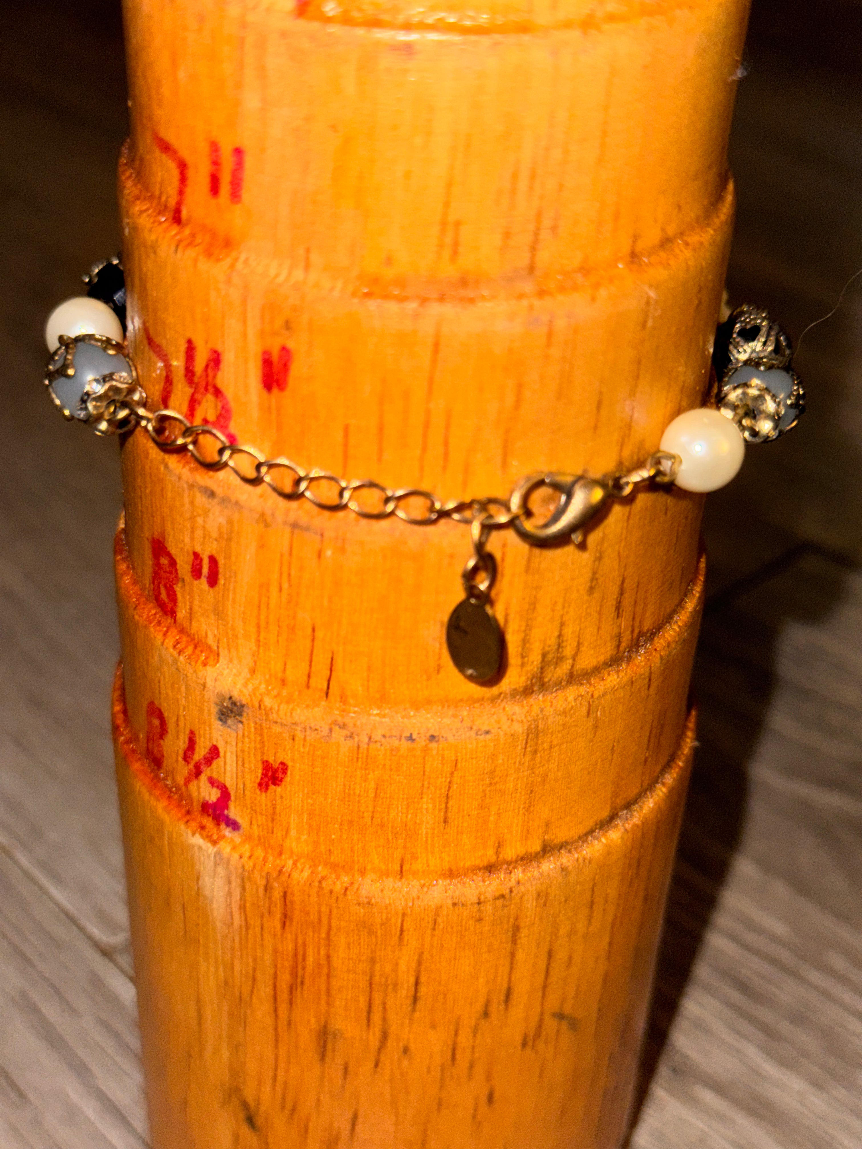 A gold charm bracelet by Accessorize with white pearl-like beads and dark metallic accents, displayed on a wooden cylindrical stand.