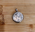 A round silver locket pendant with an embossed floral scroll pattern, resting on a light brown wooden surface.