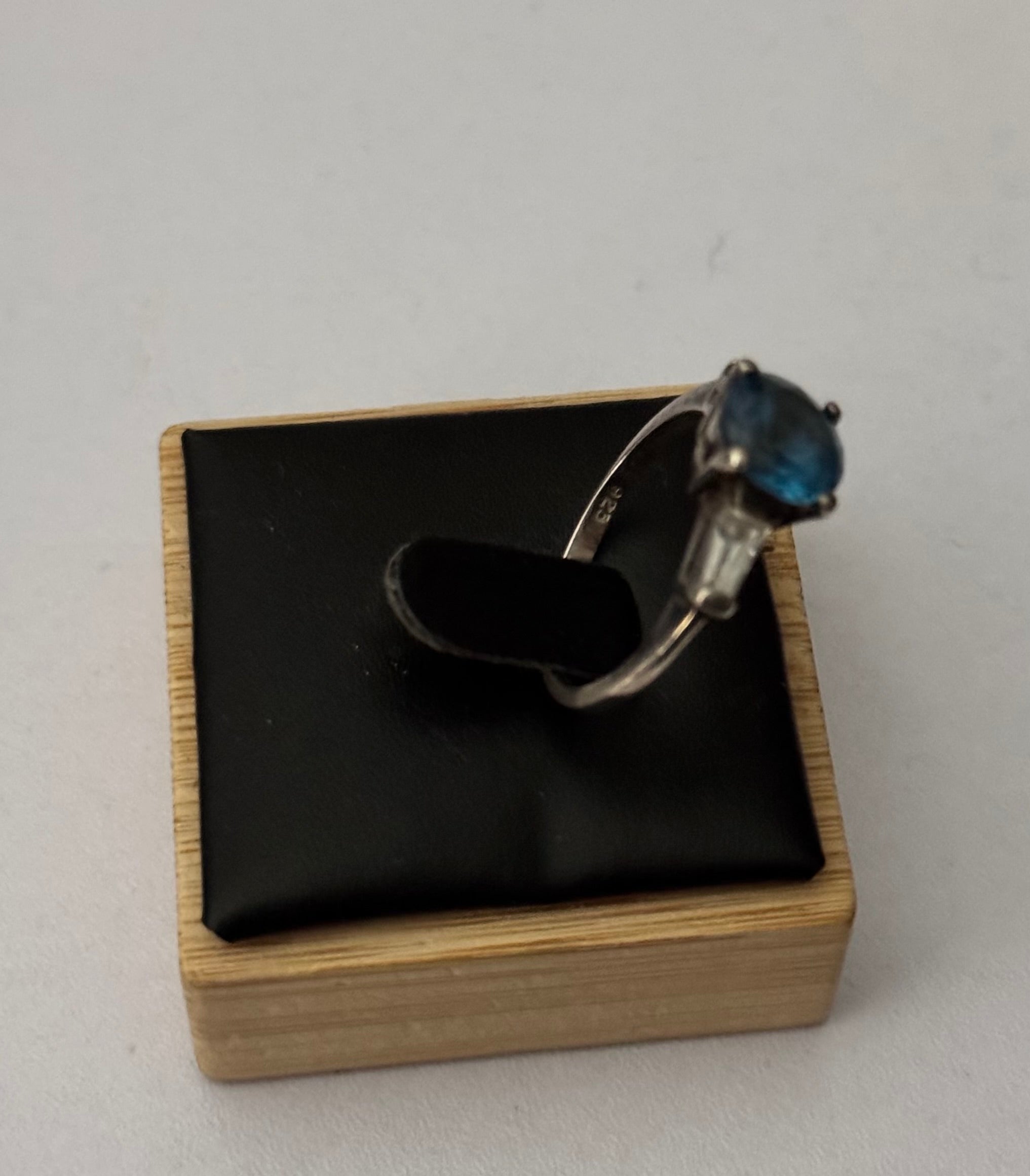 A silver ring with a blue oval gemstone believed to be a sapphire displayed on a black cushioned wooden jewelry box against a light gray background. From the side.