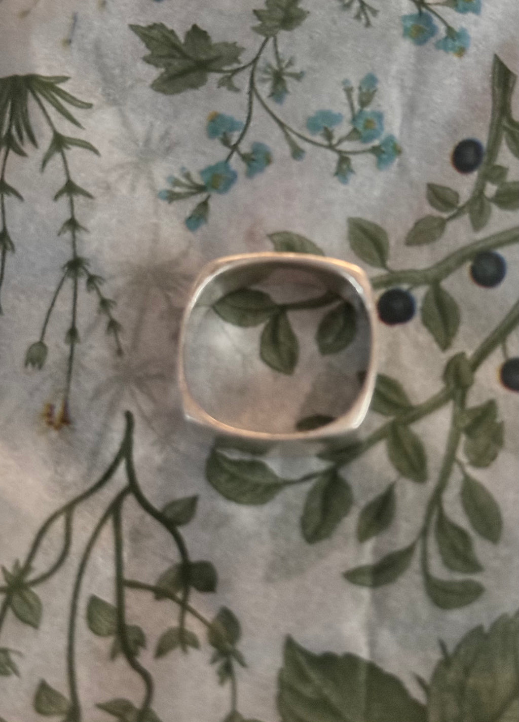 A sterling silver band ring with a square exterior & round center, resting on a light gray fabric background with a green and blue botanical print.
