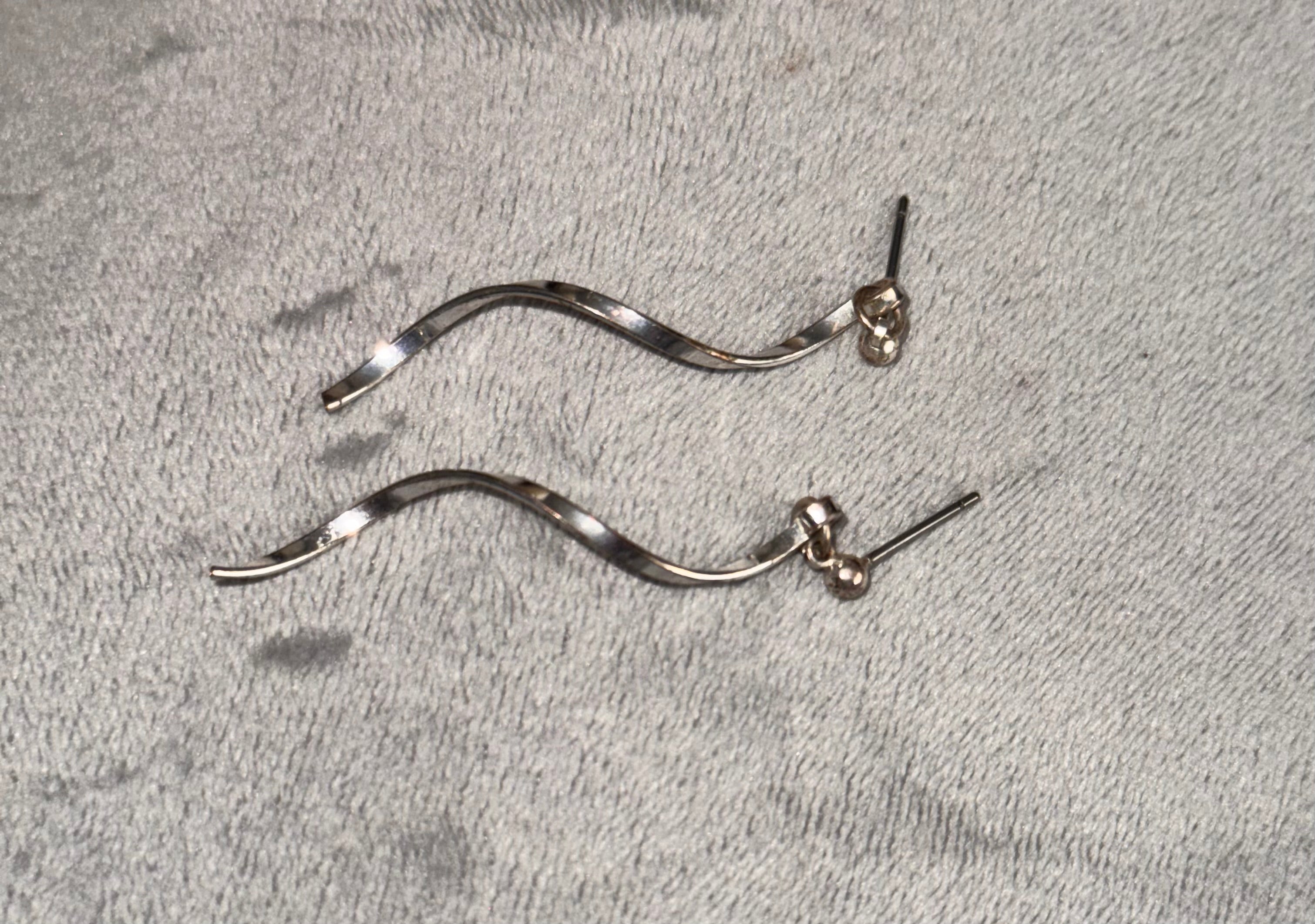 A pair of silver wavy drop earrings with small bead details and post backs, displayed on a textured gray fabric background.