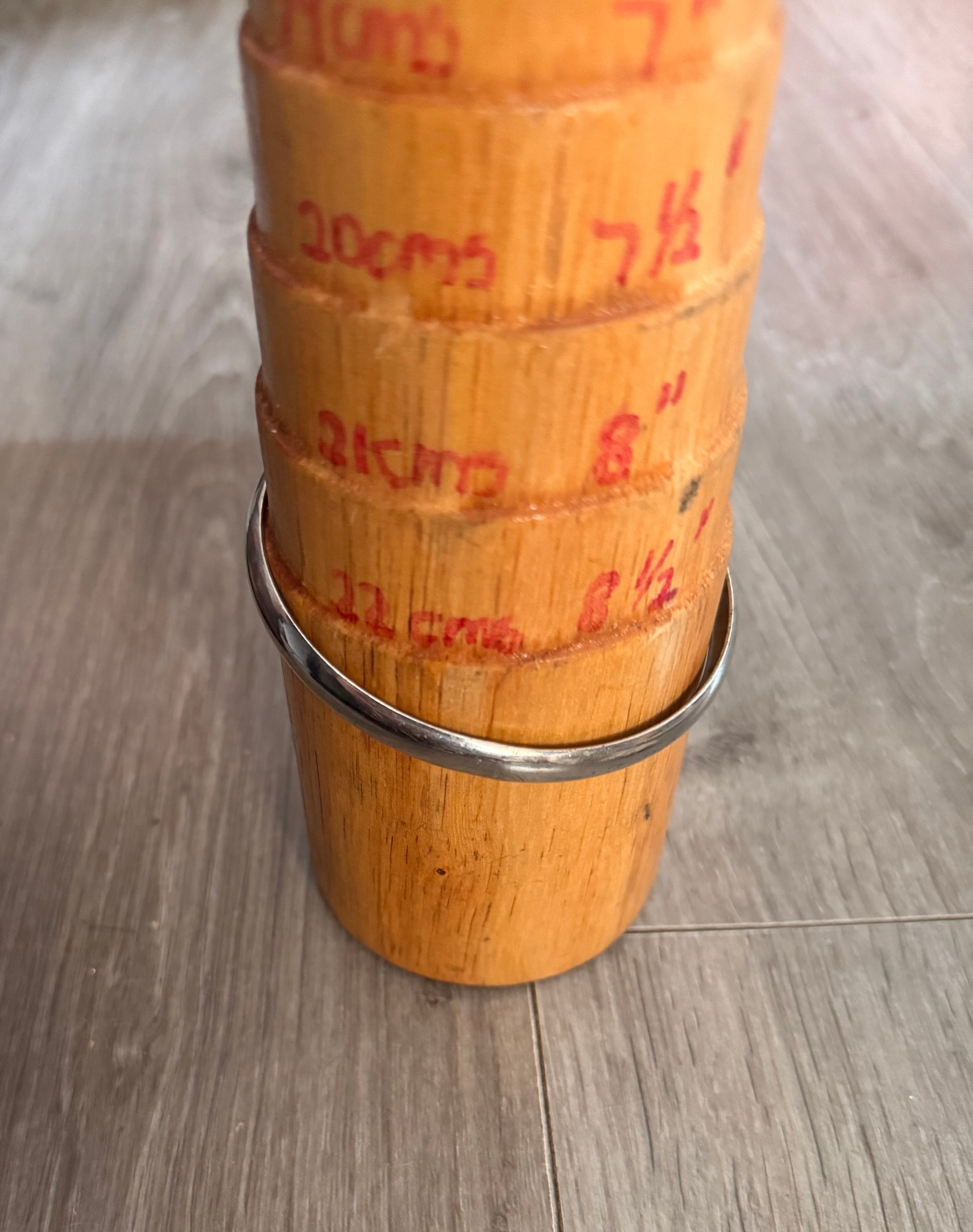 A bracelet mandrel with red handwritten measurements, displaying a silver toned non precious metal bangle, on a gray wood-look floor.