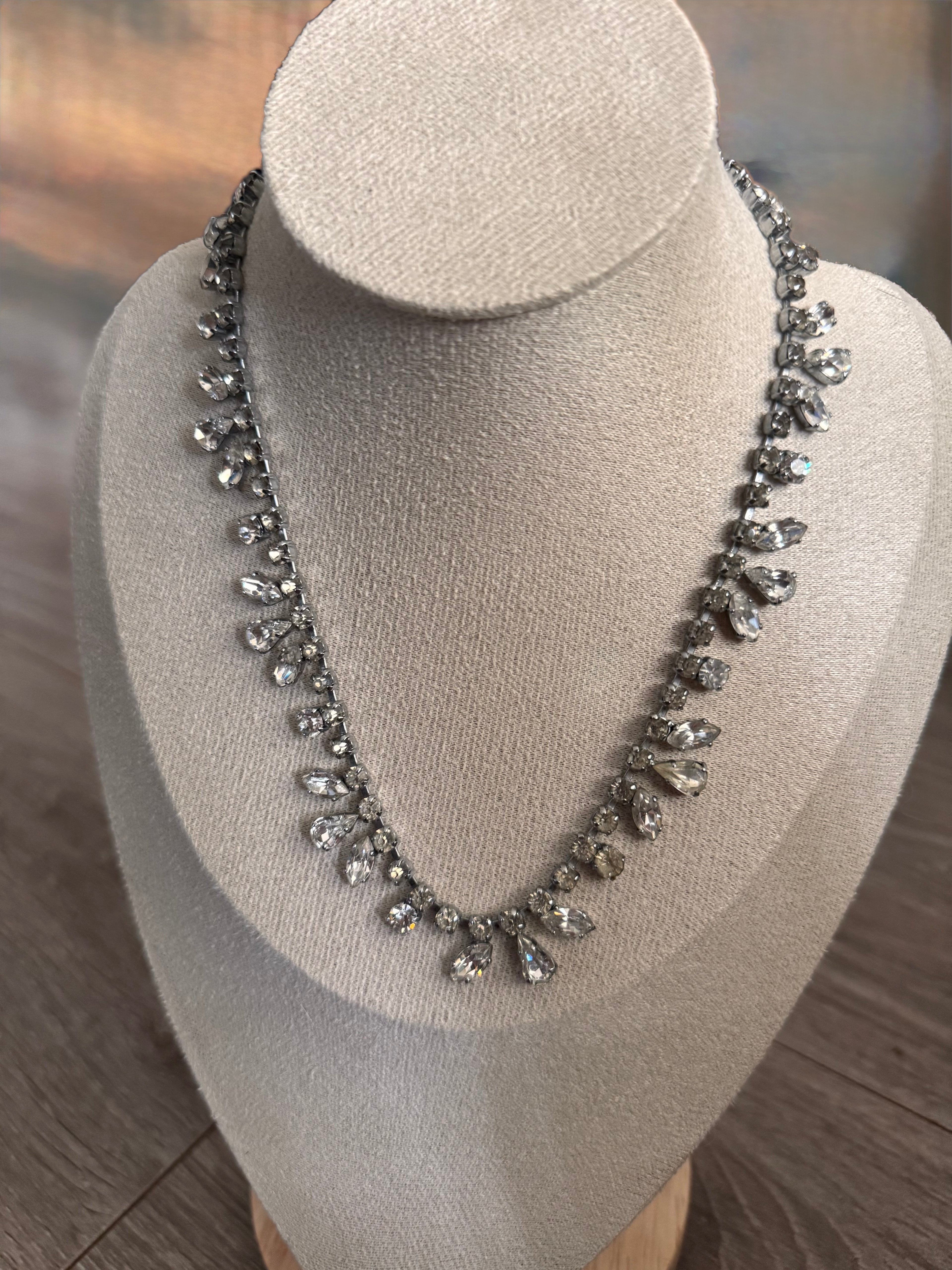 A silver-toned teardrop crystal necklace displayed on a beige fabric jewelry bust against a blurred wood background.