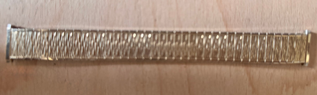 A close-up of a metallic watch band, good gold plate, with a textured, ribbed pattern, shown horizontally on a light beige background.