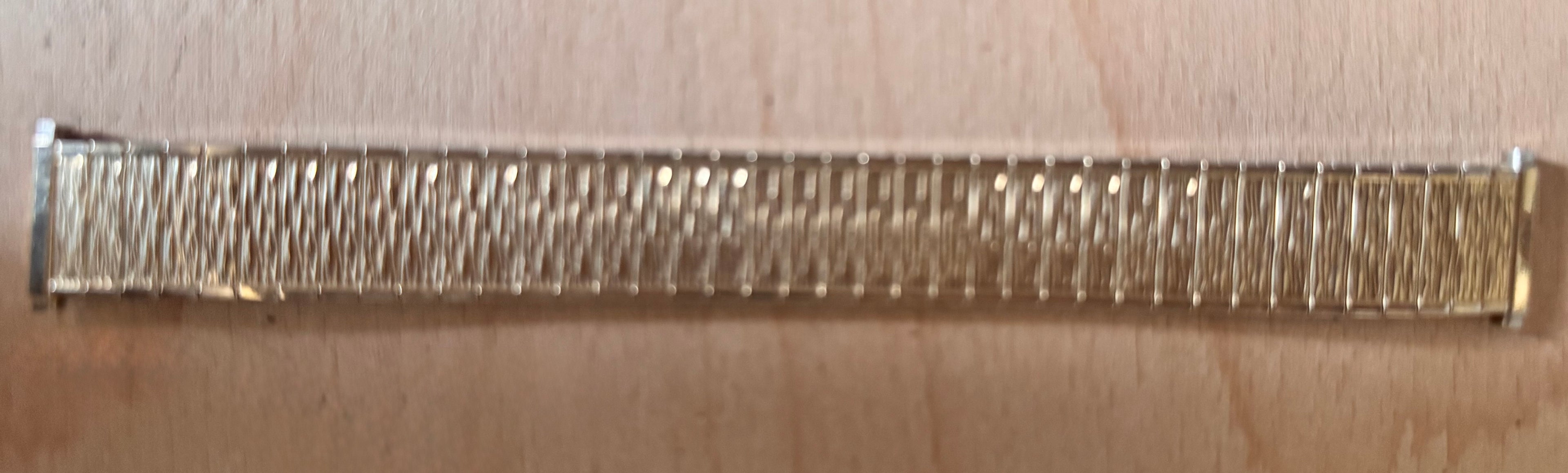 A close-up of a metallic watch band, good gold plate, with a textured, ribbed pattern, shown horizontally on a light beige background.