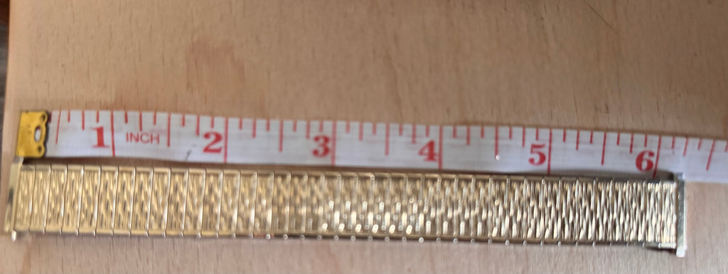 A close-up of a gold-toned metal link band, likely a watch band or bracelet, shown horizontally on a light wood surface with a white measuring tape ruler above it for scale.