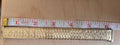 A close-up of a gold-toned metal link band, likely a watch band or bracelet, shown horizontally on a light wood surface with a white measuring tape ruler above it for scale.