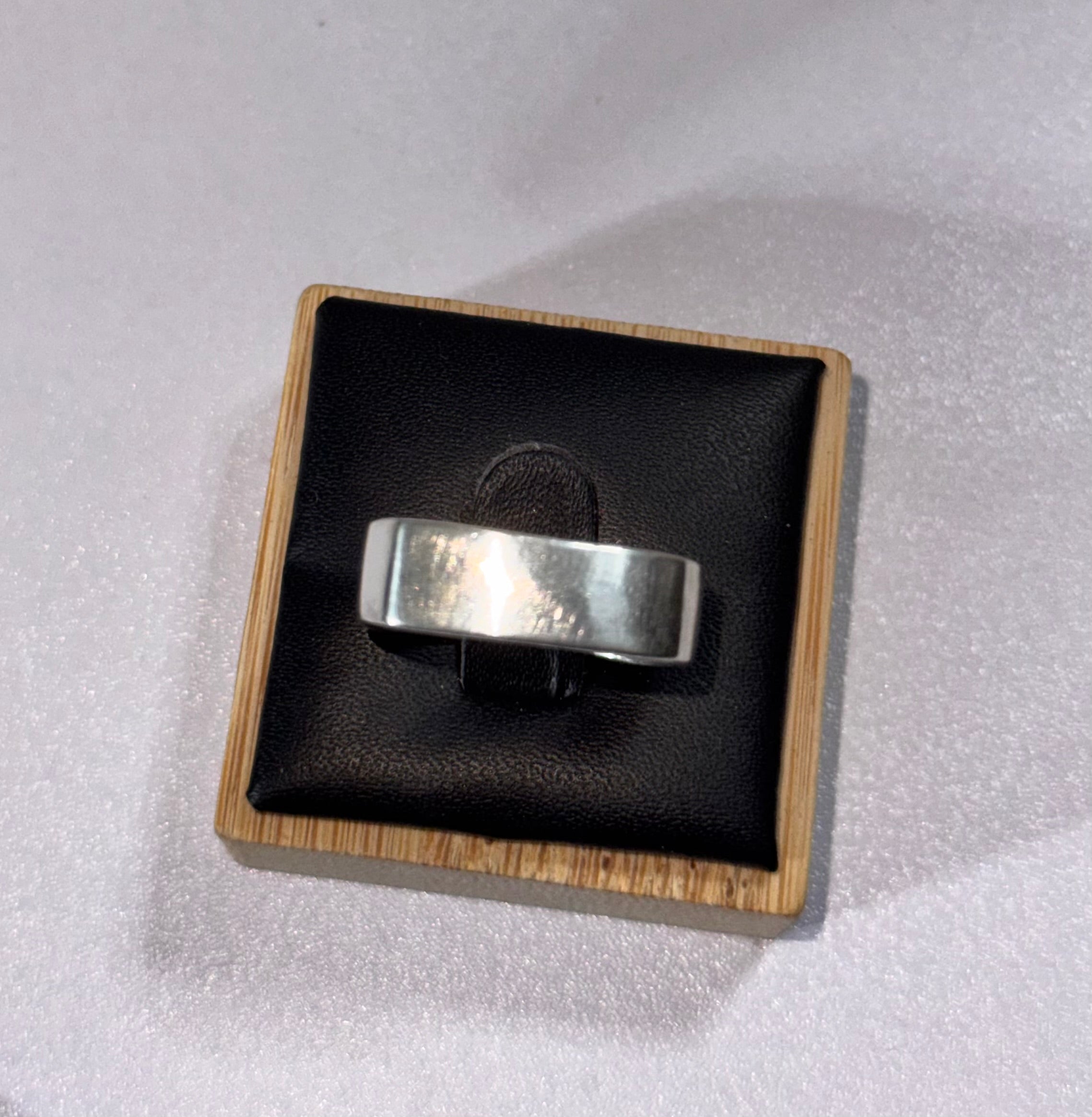 A sterling silver band ring displayed on a black cushion inside a small square wooden presentation box, set against a light gray textured background.