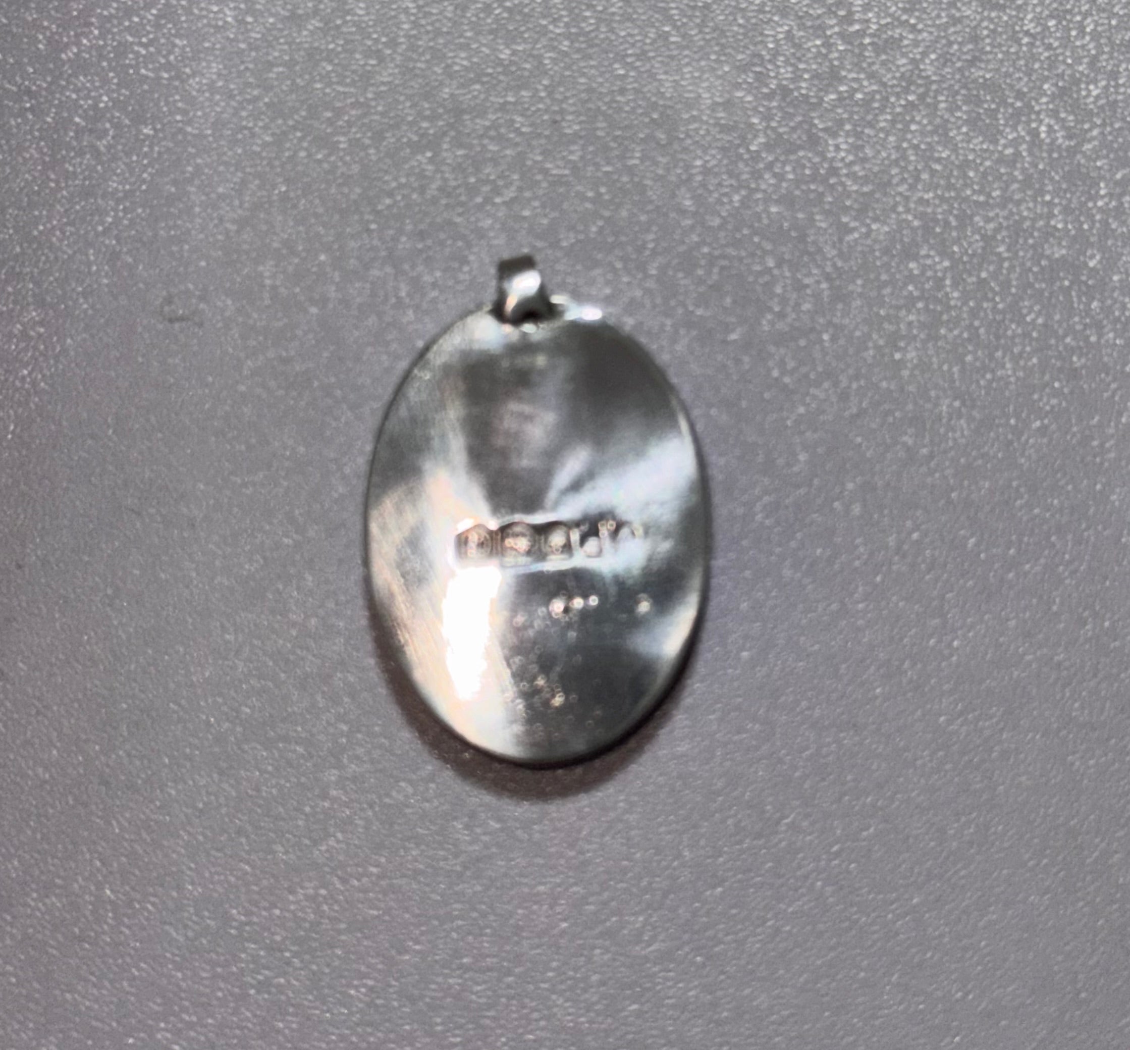 A 2cm length oval sterling silver pendant with a bail at the top, photographed on a textured gray background. Displaying the original hallmark
