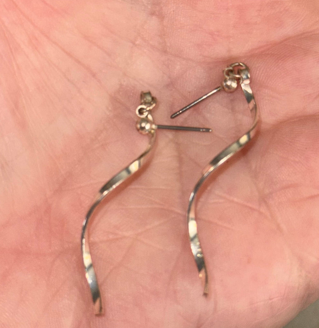 A pair of dangling spiral earrings with a smooth metallic finish, resting on an open palm. The earrings appear to be made of a warm-toned silver plated metal alloy, with flat post backings.