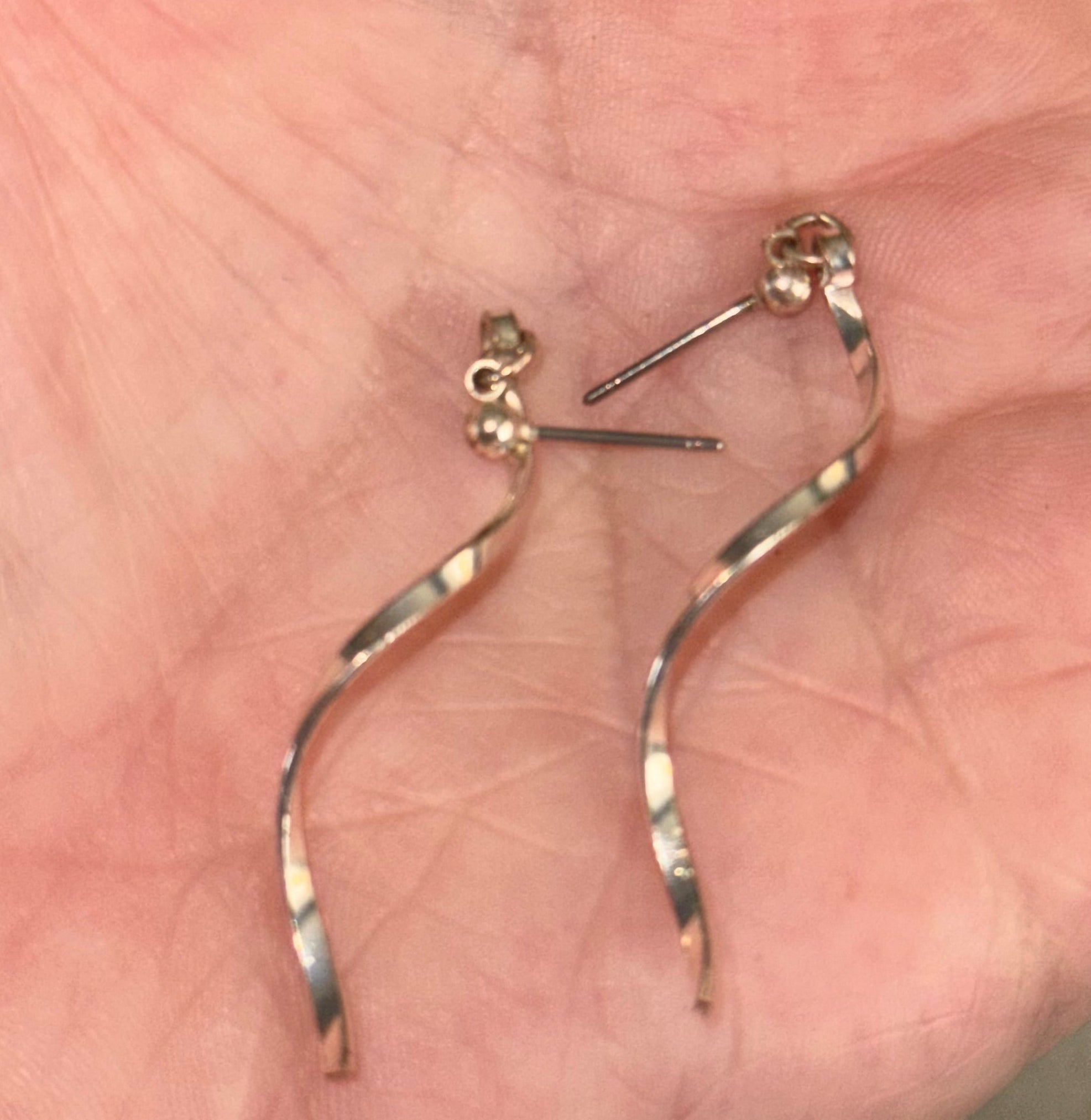 A pair of dangling spiral earrings with a smooth metallic finish, resting on an open palm. The earrings appear to be made of a warm-toned silver plated metal alloy, with flat post backings.