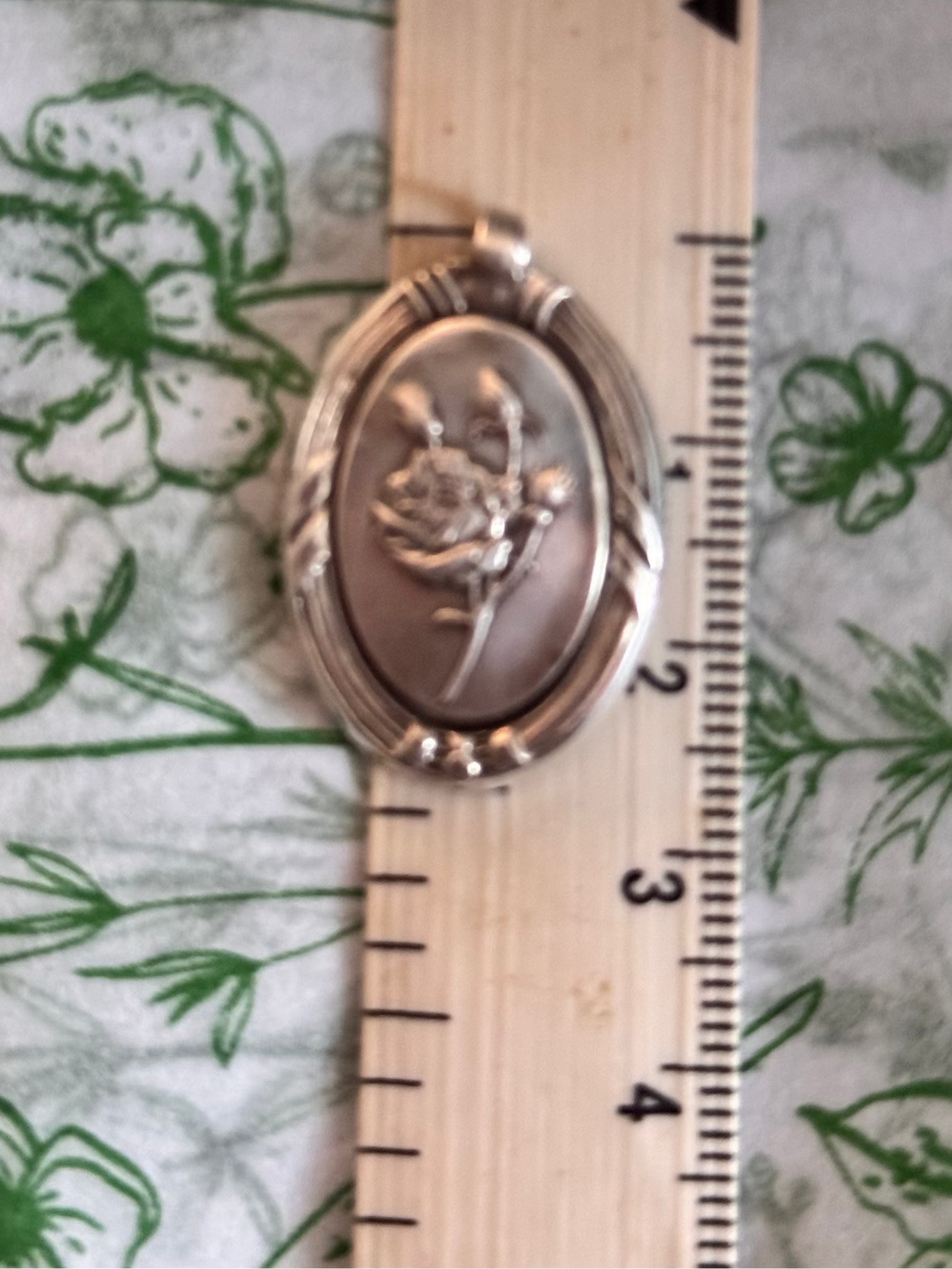 A sterling silver oval pendant with a Rose design, shown next to a measure showing the height of 2.6cm on a green floral-patterned fabric background.