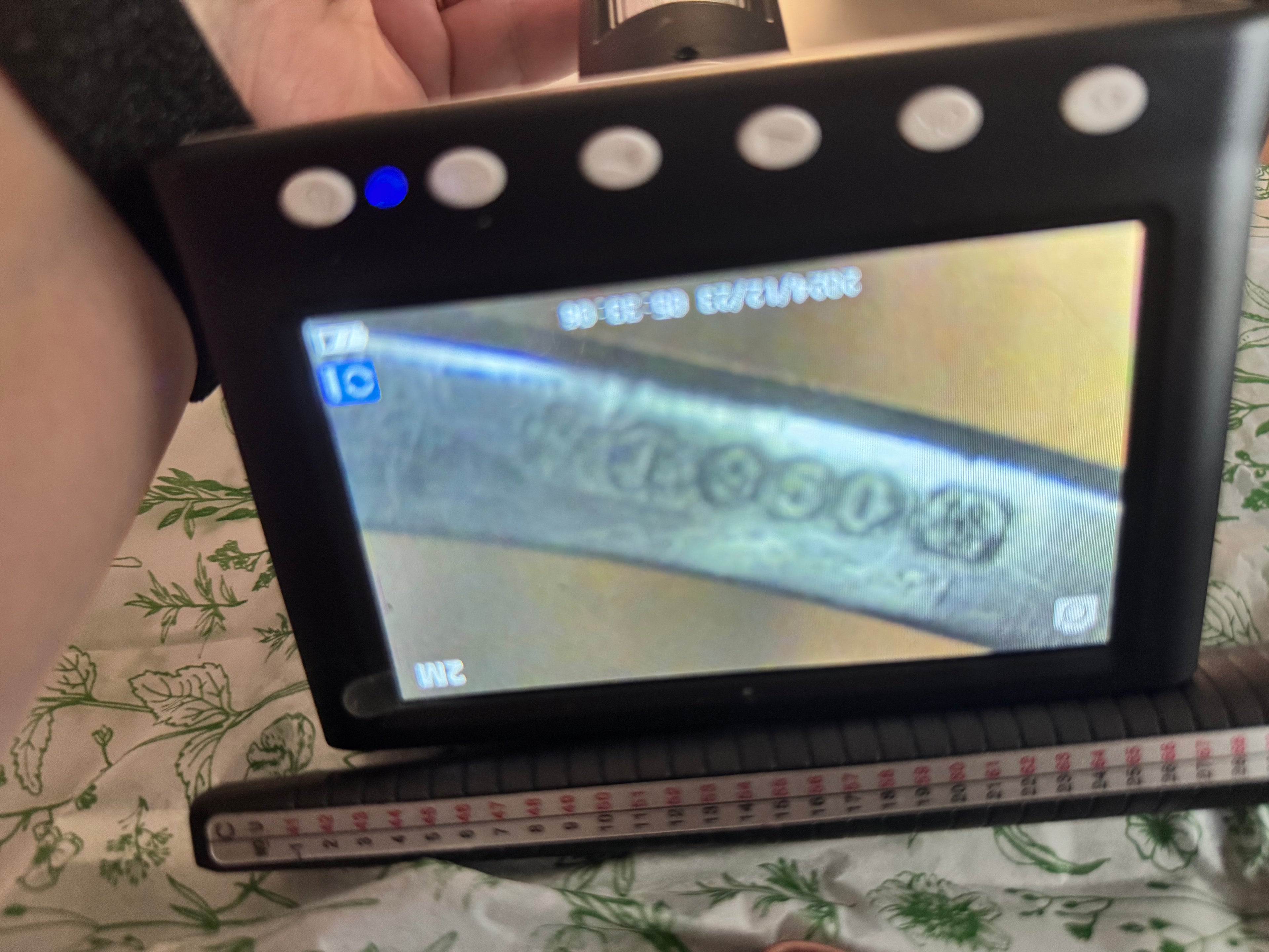 A handheld black inspection microscope or monitor with a screen showing a close-up of engraved hallmark on the ring, held by a person over a patterned green and white fabric surface with a ring guage in front.