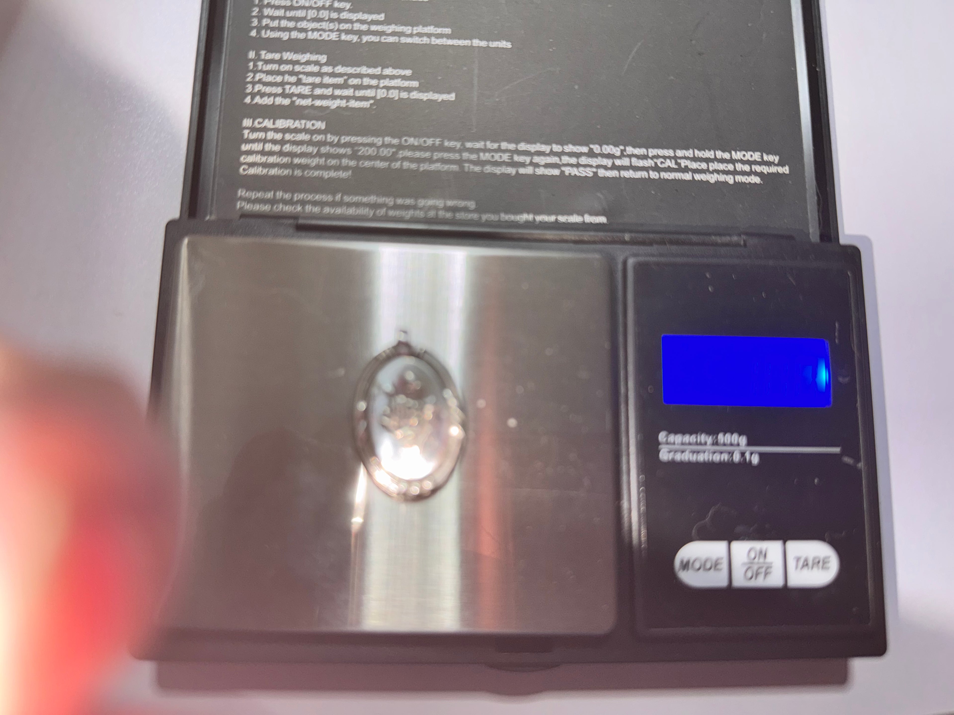 A close-up of a digital pocket scale  showing 10 grams but difficult to see.  The pendant is sat on a stainless steel weighing platform and a blue display screen showing blue screen display. 