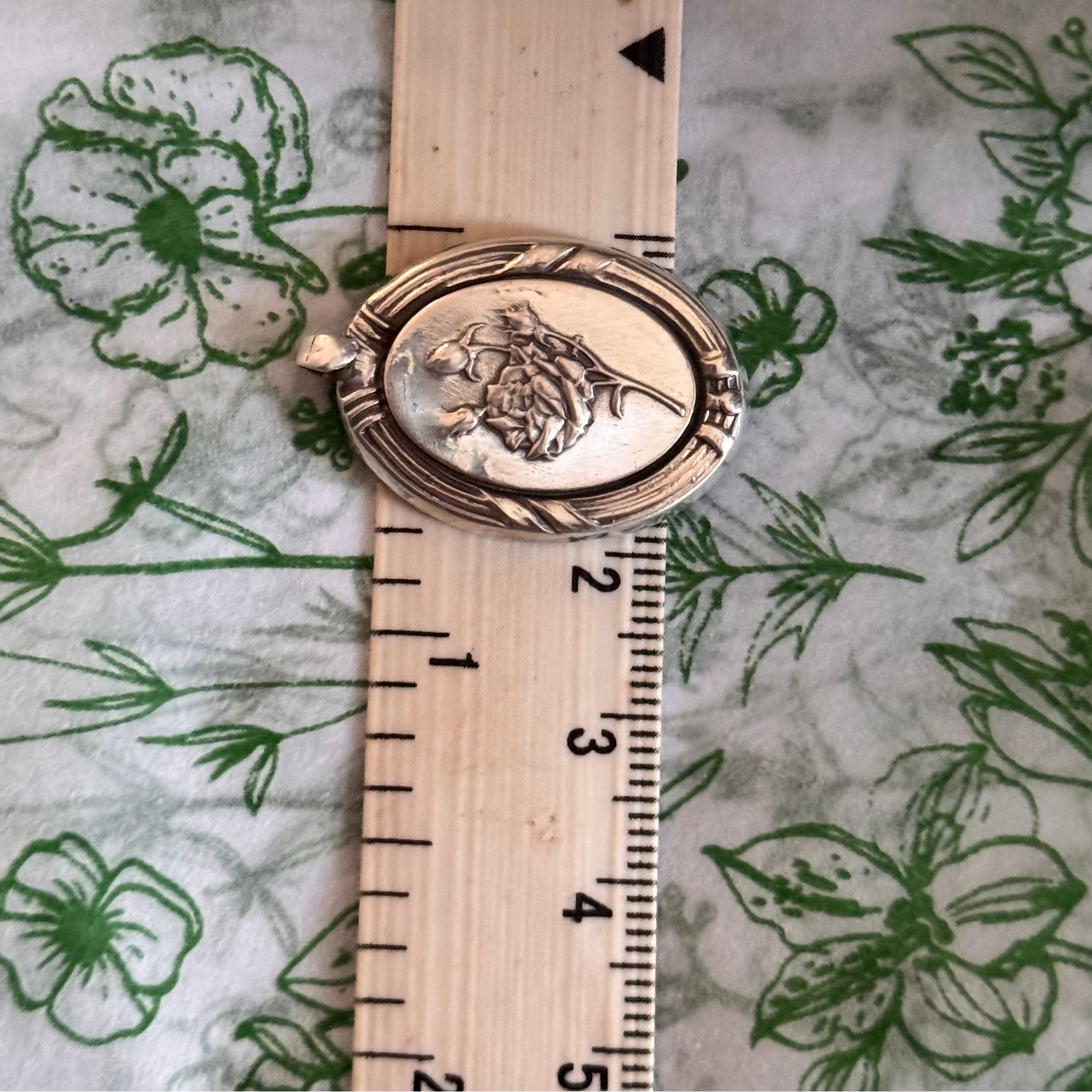 The sterling Silver rose pendant on a measuring tape. on a background of Floral green and white material. Shows the width of 2cm