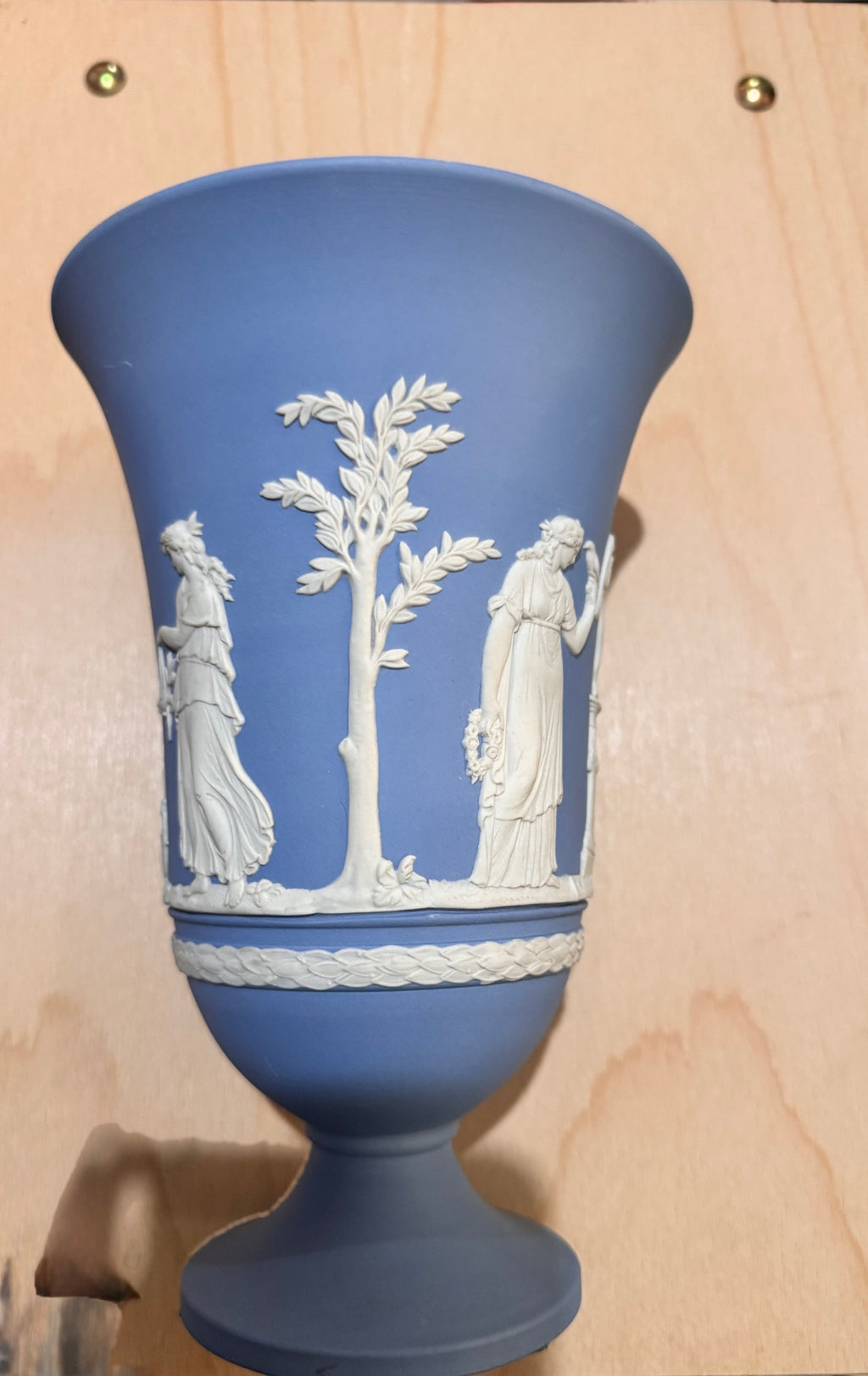 A blue and white Wedgwood ceramic pedestal Vase with a relief band depicting classical figures and a tree, shown against a light wood background. Vintage Blue Jasperware by Wedgewood
