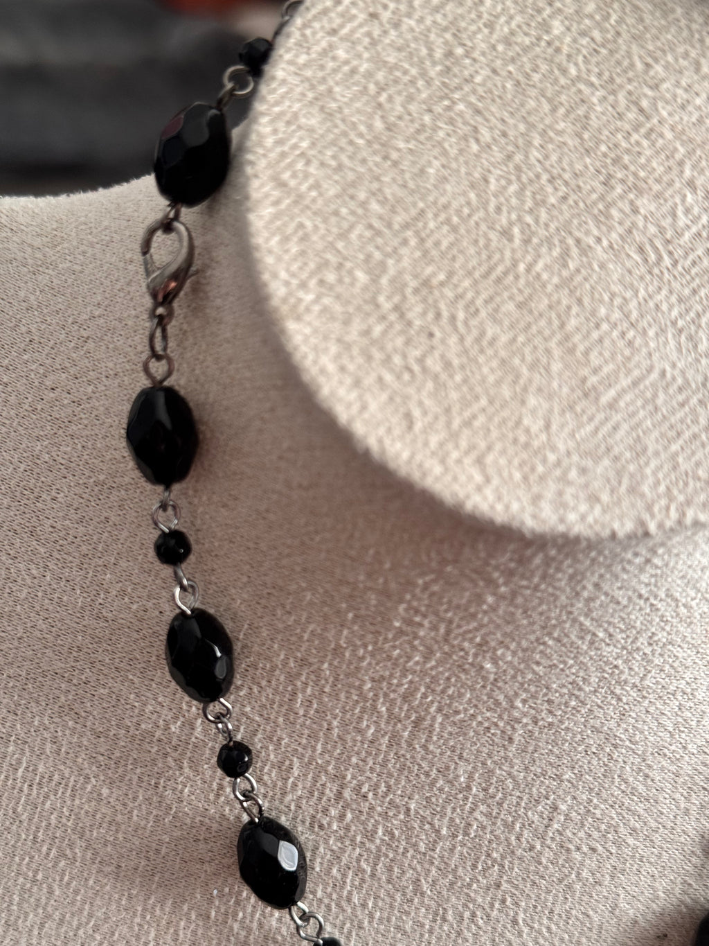 A close-up of a beaded necklace with faceted black beads and small black spacer beads on a silver chain, displayed on a textured beige fabric jewelry bust. Displaying the lobster clasp too.