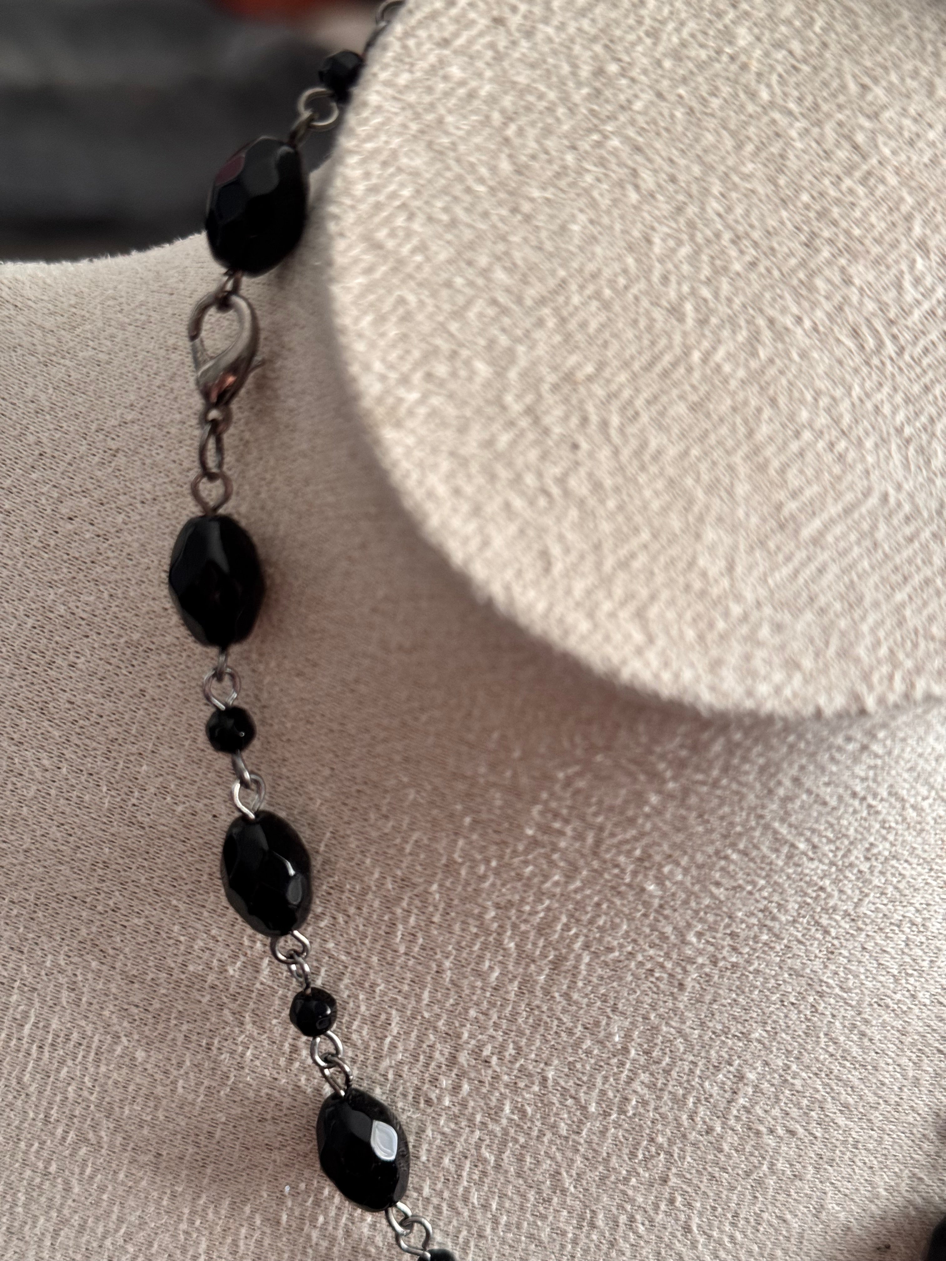 A close-up of a beaded necklace with faceted black beads and small black spacer beads on a silver chain, displayed on a textured beige fabric jewelry bust. Displaying the lobster clasp too.