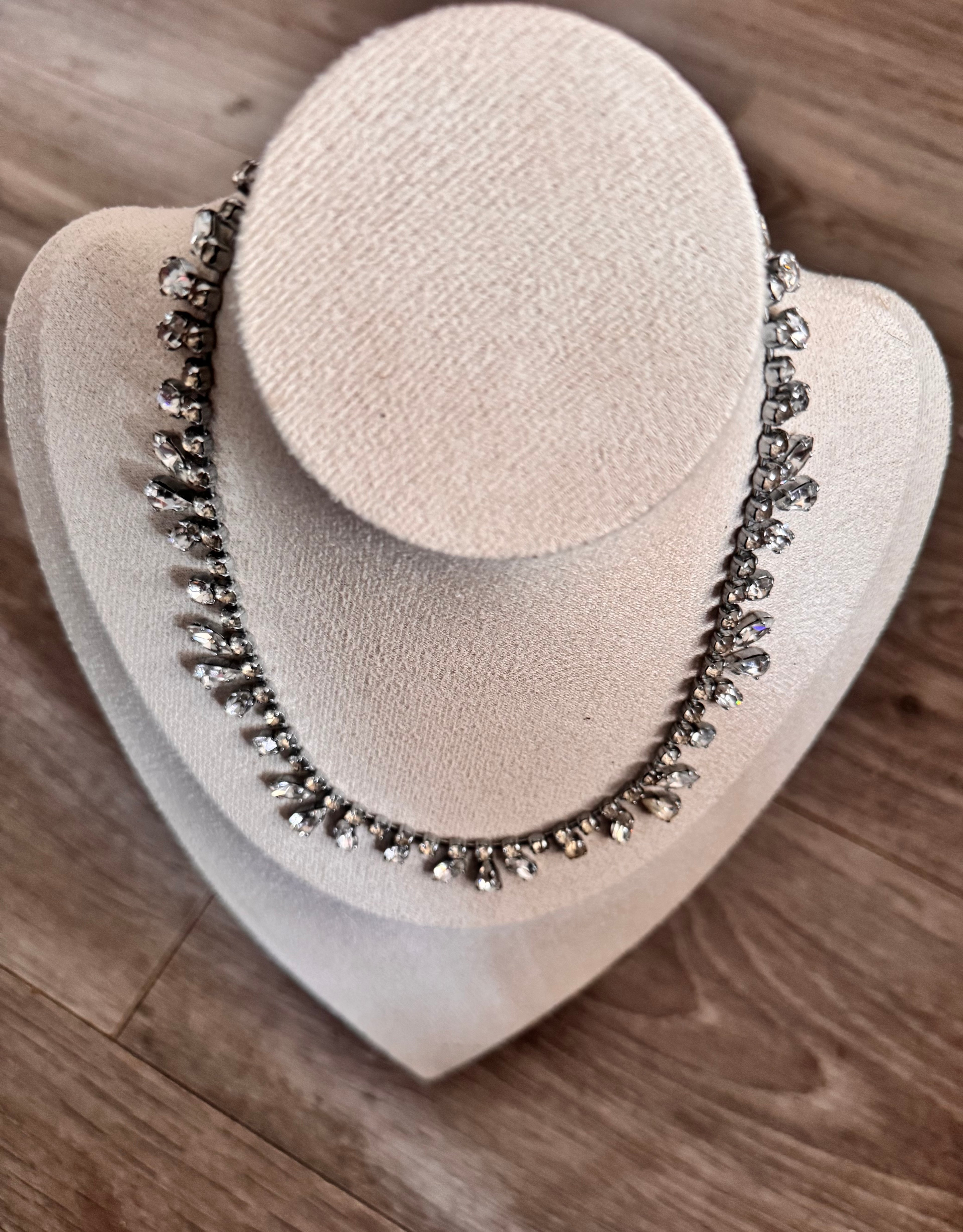 A silver-toned beaded necklace with clear rhinestone-like accents, displayed on a cream-colored jewelry bust against a wood floor background.