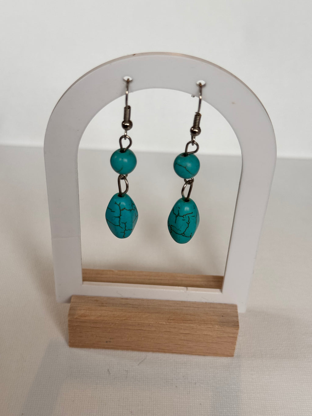 A pair of turquoise drop earrings with silver-toned metal hooks, displayed on a white and light wood hanger against a white wall.