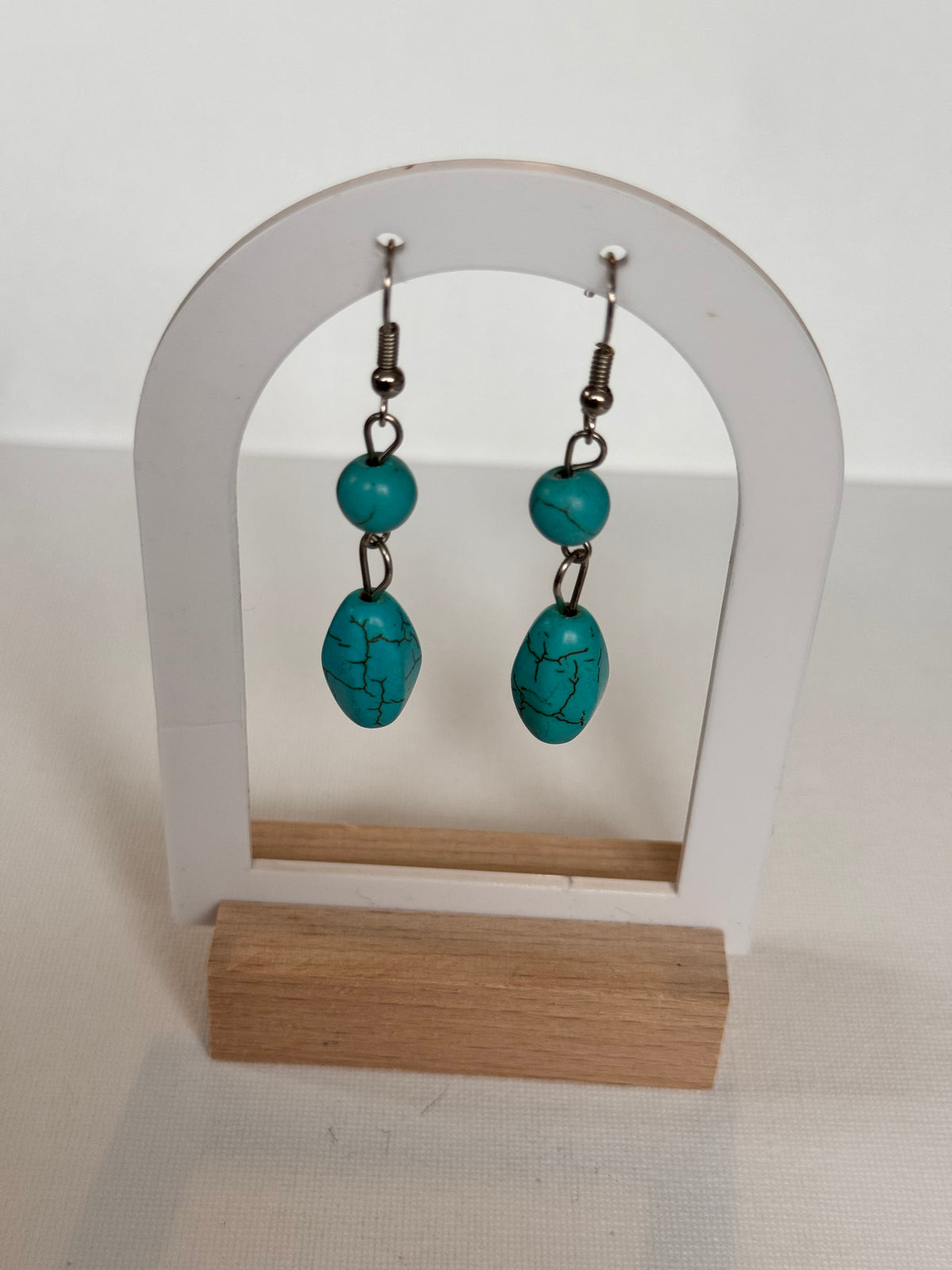 A pair of turquoise drop earrings with silver-toned metal hooks, displayed on a white and light wood hanger against a white wall.