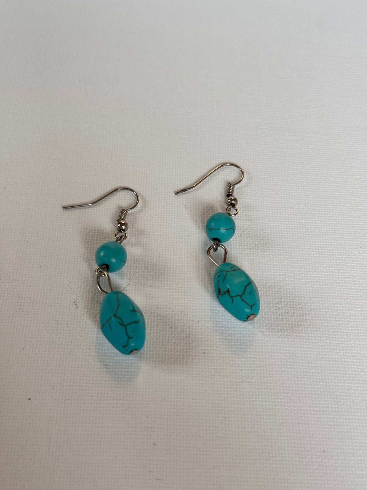 A pair of dangle earrings featuring turquoise-blue marbled beads and small round turquoise beads on silver fishhook wires, displayed on a textured off-white fabric background.