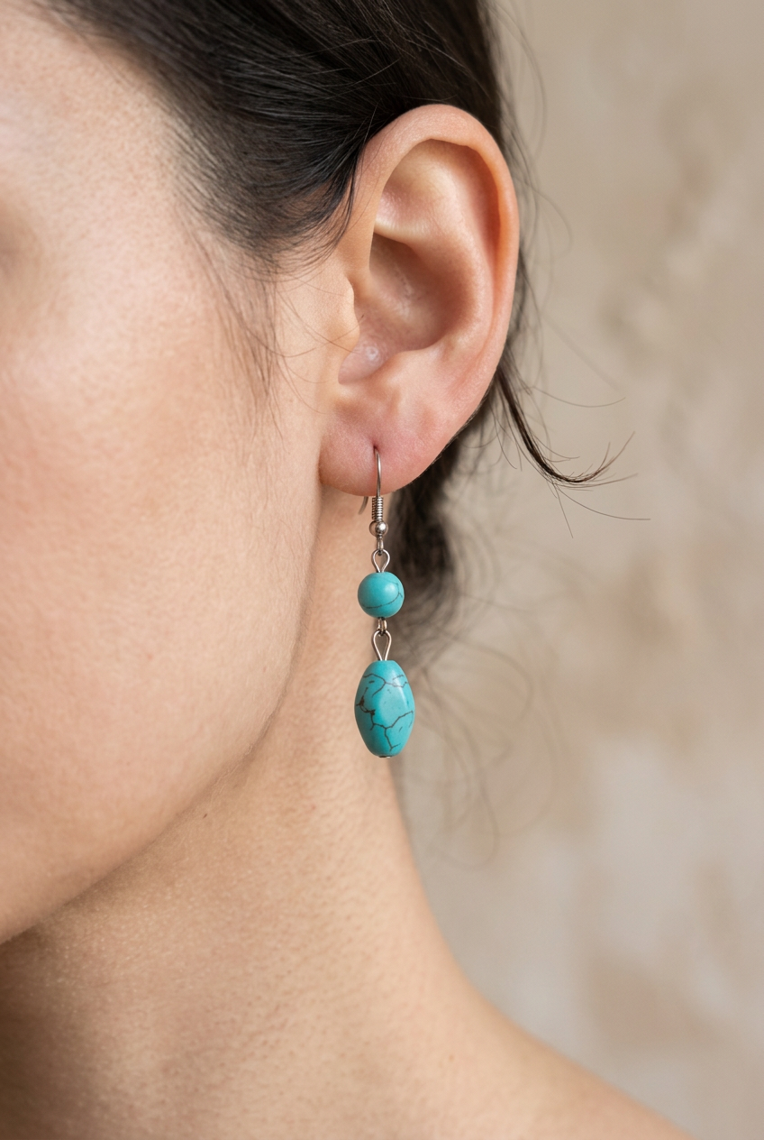 A close-up of a model's ear wearing a double-drop dangle earring. The earring features a small round turquoise bead above a larger, irregular turquoise stone set in silver-toned metal. This is an AI image to show how they look in the ear. The other images are true and not AI