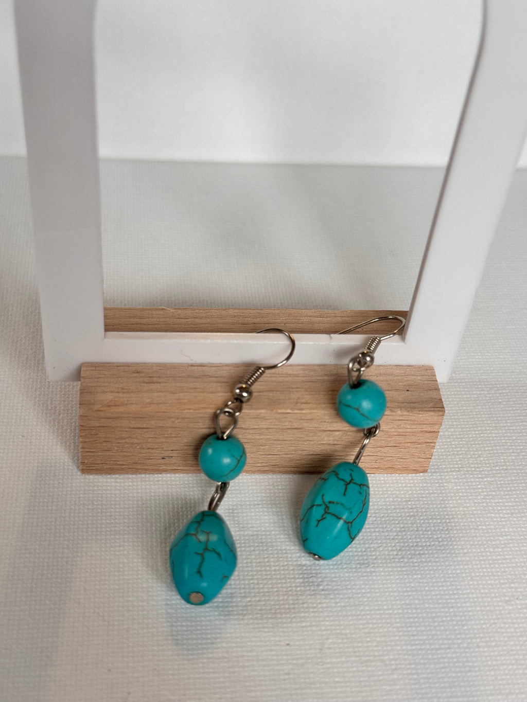 A pair of turquoise drop earrings with silver hooks and small round turquoise beads, hanging from a wooden display stand against a white textured background.