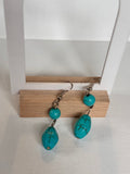 A pair of turquoise drop earrings with silver hooks and small round turquoise beads, hanging from a wooden display stand against a white textured background.