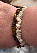 Two beaded stretch bracelets worn on a wrist. One bracelet features polished brown tiger's eye stones, and the other features smooth white river stones.