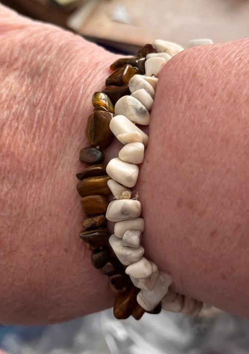 Two beaded stretch bracelets worn on a wrist. One bracelet features polished brown tiger's eye stones, and the other features smooth white river stones.