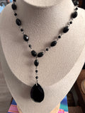 A beaded necklace with black faceted beads and a large black teardrop pendant, displayed on a beige jewelry bust.