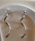 A pair of silver-toned wavy dangle earrings displayed on a white arch stand.